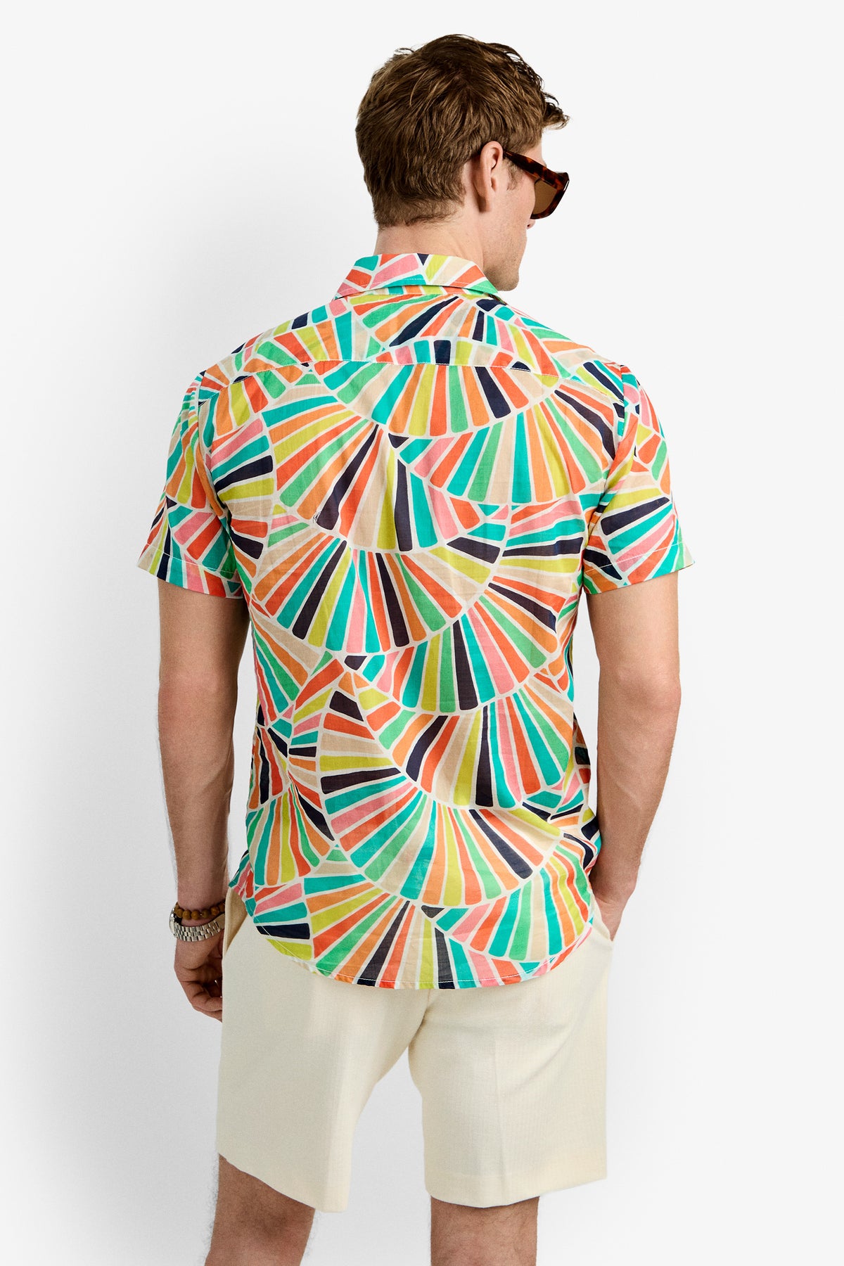 A man wearing sunglasses and a Slim Jim Shirt with a colorful geometric pattern stands in light-colored shorts facing away from the camera against a plain white background.