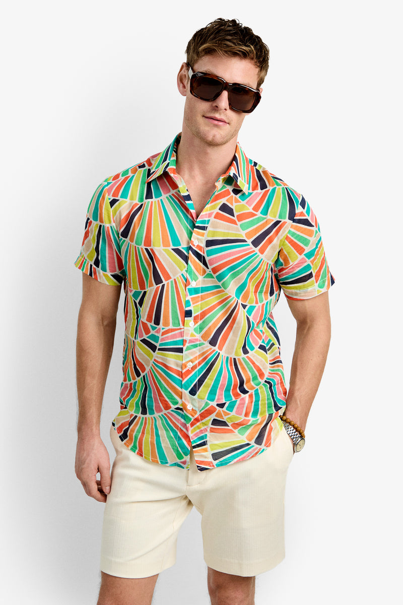 A man wearing the Slim Jim Shirt—a colorful short-sleeve button-up with a geometric pattern—large dark sunglasses, and light beige shorts stands against a plain white background.