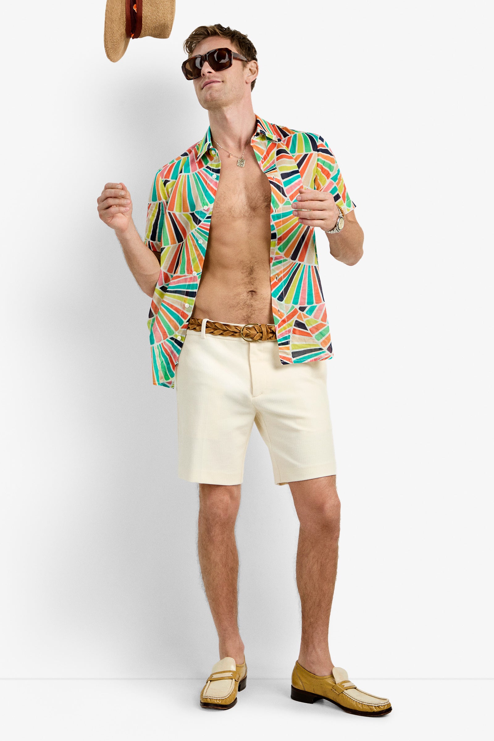 A man wearing sunglasses, the Slim Jim Shirt in Coppelia (unbuttoned), cream shorts, a braided belt, and two-tone loafers stands against a white background, tossing a straw hat into the air.