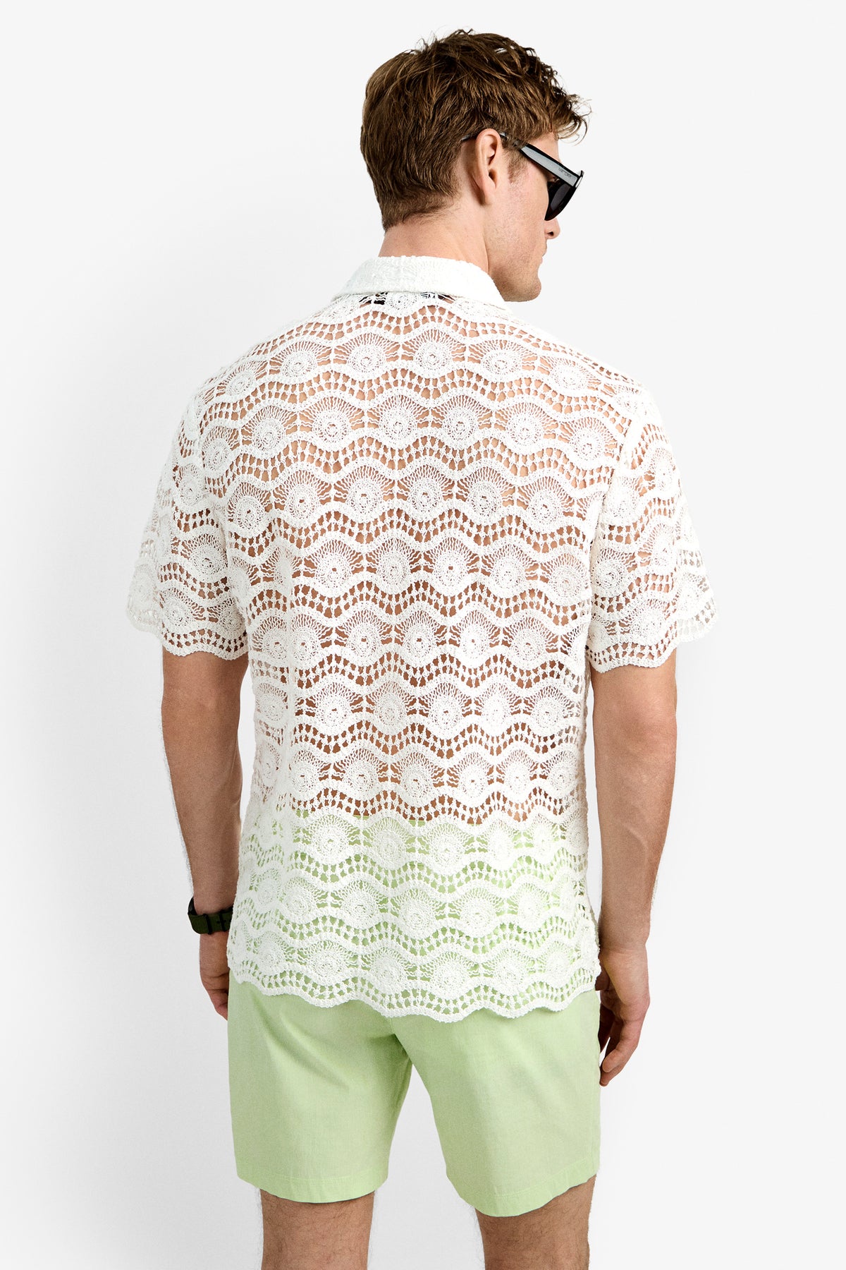 A man faces away from the camera against a plain white background, wearing black sunglasses, light green shorts, and the Long Beach Shirt, a white short-sleeve crochet top.