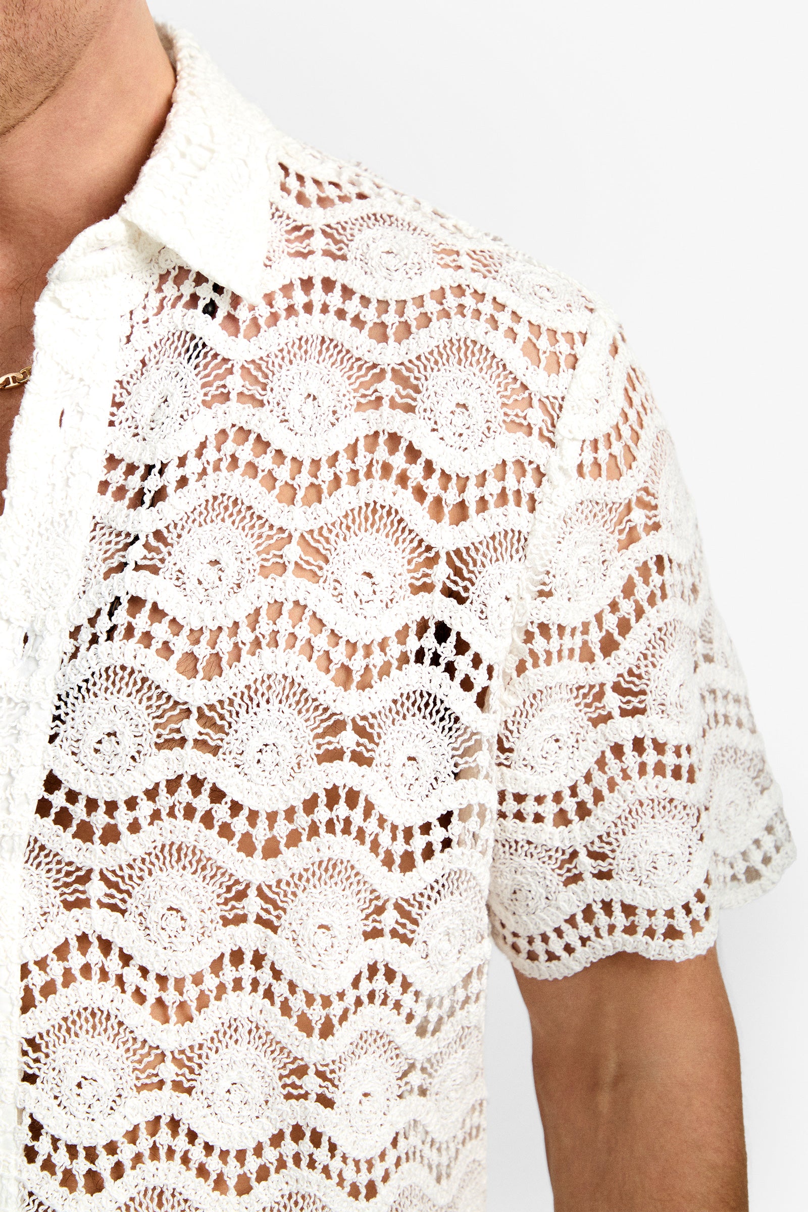 A person wearing the Long Beach Shirt, a short-sleeve white crocheted lace top with a wavy scalloped pattern. The see-through design reveals their skin beneath. Only the upper torso and part of their face are visible.