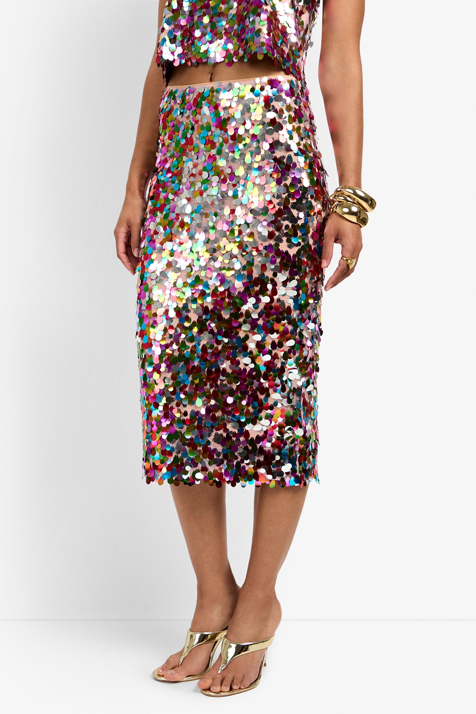 A person wearing the Pearla Skirt—a shiny, multicolored sequin skirt—paired with a matching cropped top, gold bracelets, and gold heeled sandals, stands against a plain white background.