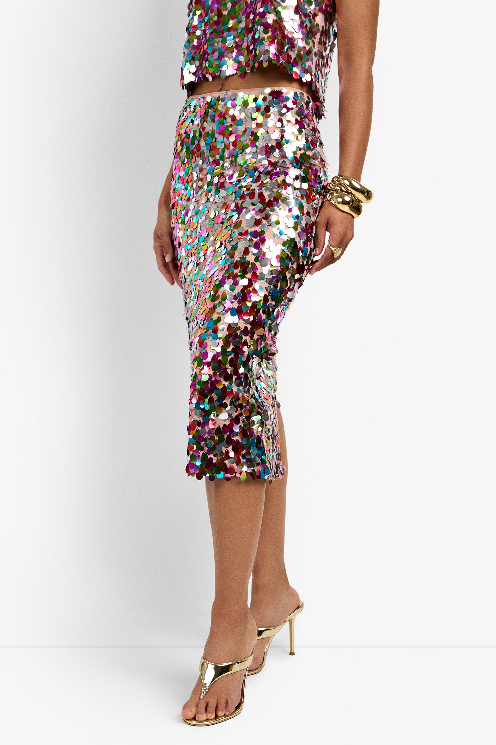 A person wearing the Pearla Skirt—a shimmering, multicolored sequin midi—paired with a matching top, gold heeled sandals, and chunky gold bracelets stands against a white background.