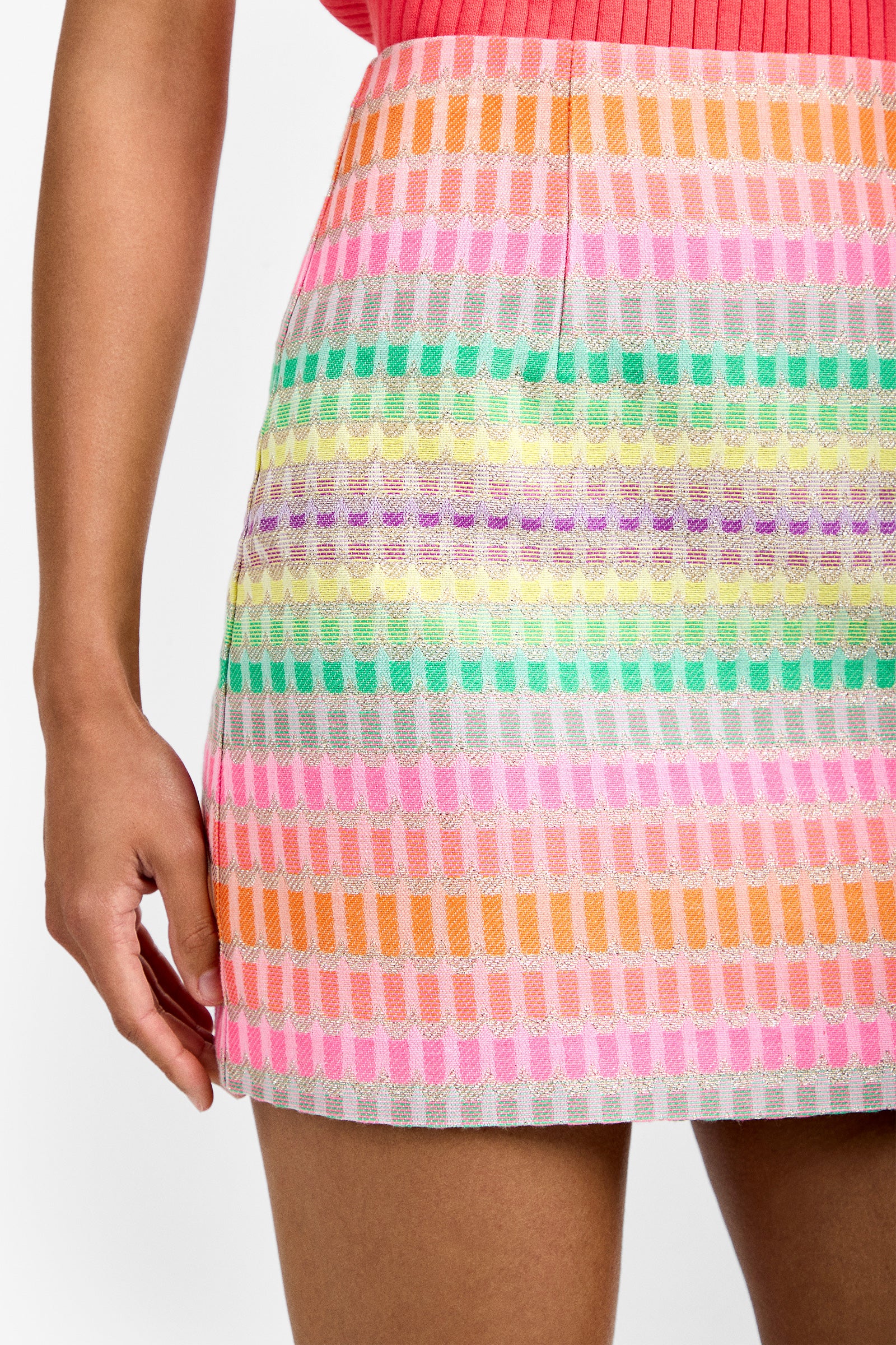 A close-up of a person wearing the High Rise Rico Skirt featuring horizontal rows of bright pastel stripes in pink, orange, green, purple, and white. Part of their arm and hand is visible on the left.