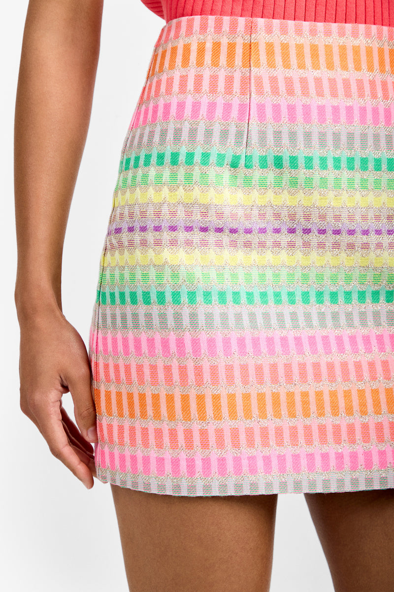 A close-up of a person wearing the High Rise Rico Skirt featuring horizontal rows of bright pastel stripes in pink, orange, green, purple, and white. Part of their arm and hand is visible on the left.