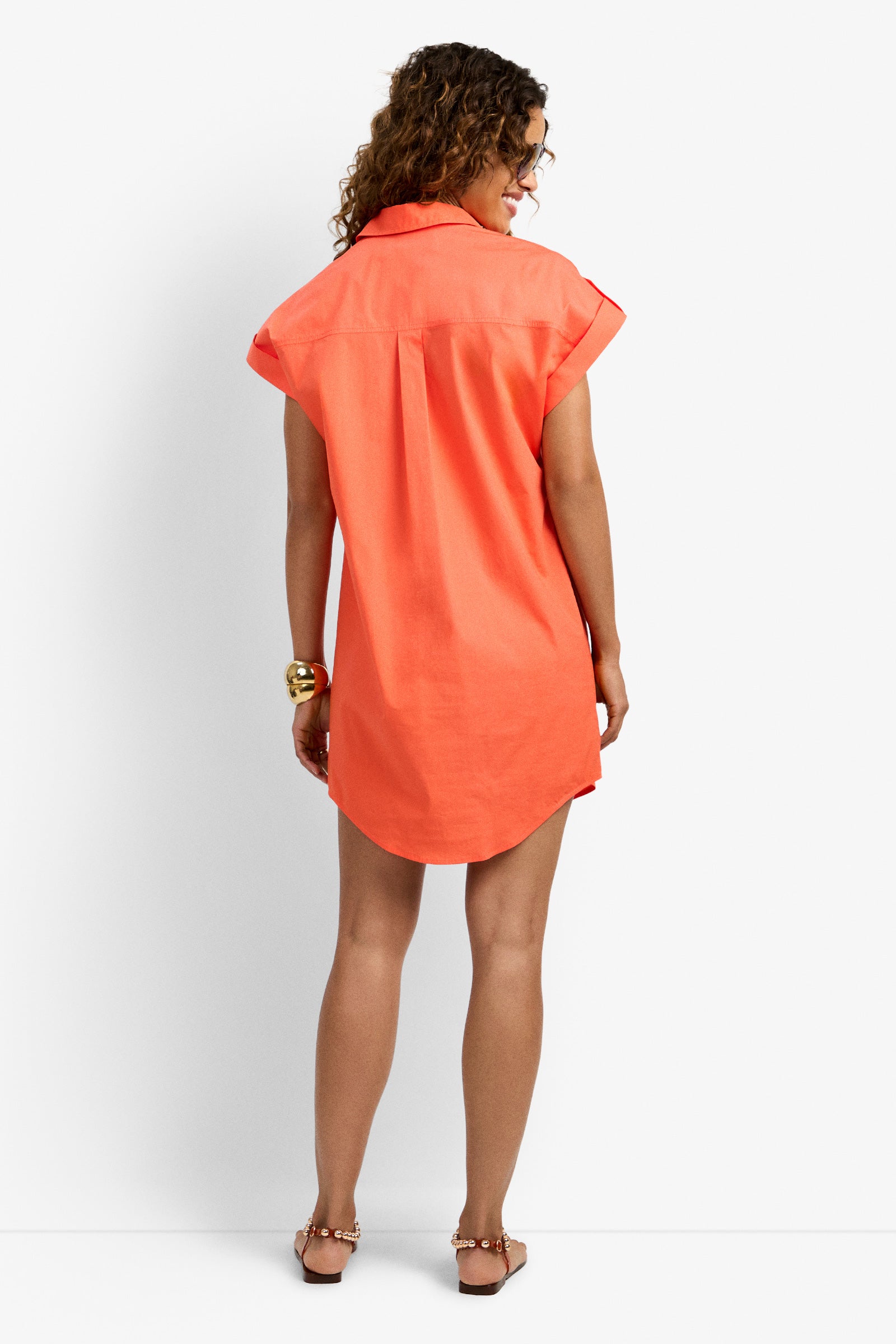 A woman with curly hair wears the Bonsoir Dress, a bright orange short-sleeve shirt dress, paired with sandals and stands with her back to the camera against a plain white background.