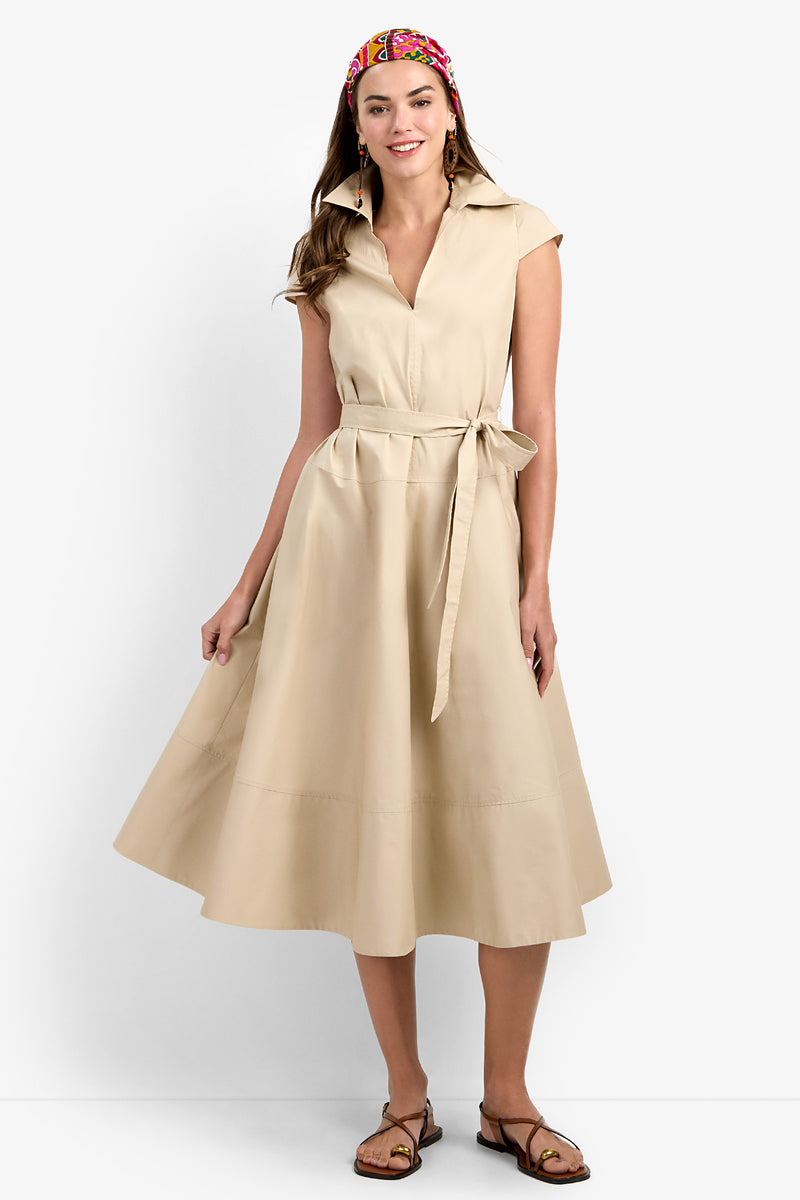 A woman stands and smiles against a plain white background, wearing the Delfina Dress—a beige, short-sleeve, collared midi dress with a waist tie—paired with brown sandals and a colorful headband.