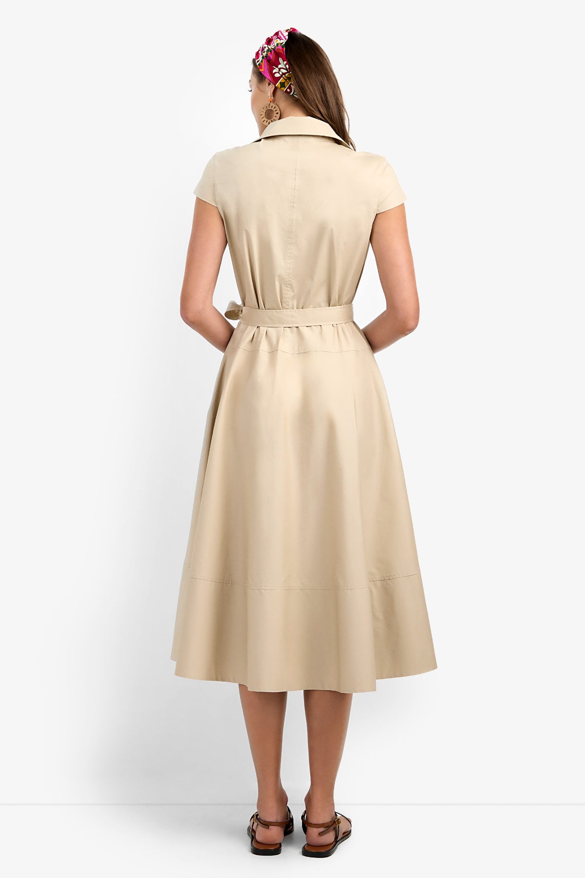 A woman faces away from the camera in a Delfina Dress—a short-sleeve beige belted midi—paired with brown sandals and a vibrant hair accessory, set against a plain white backdrop.