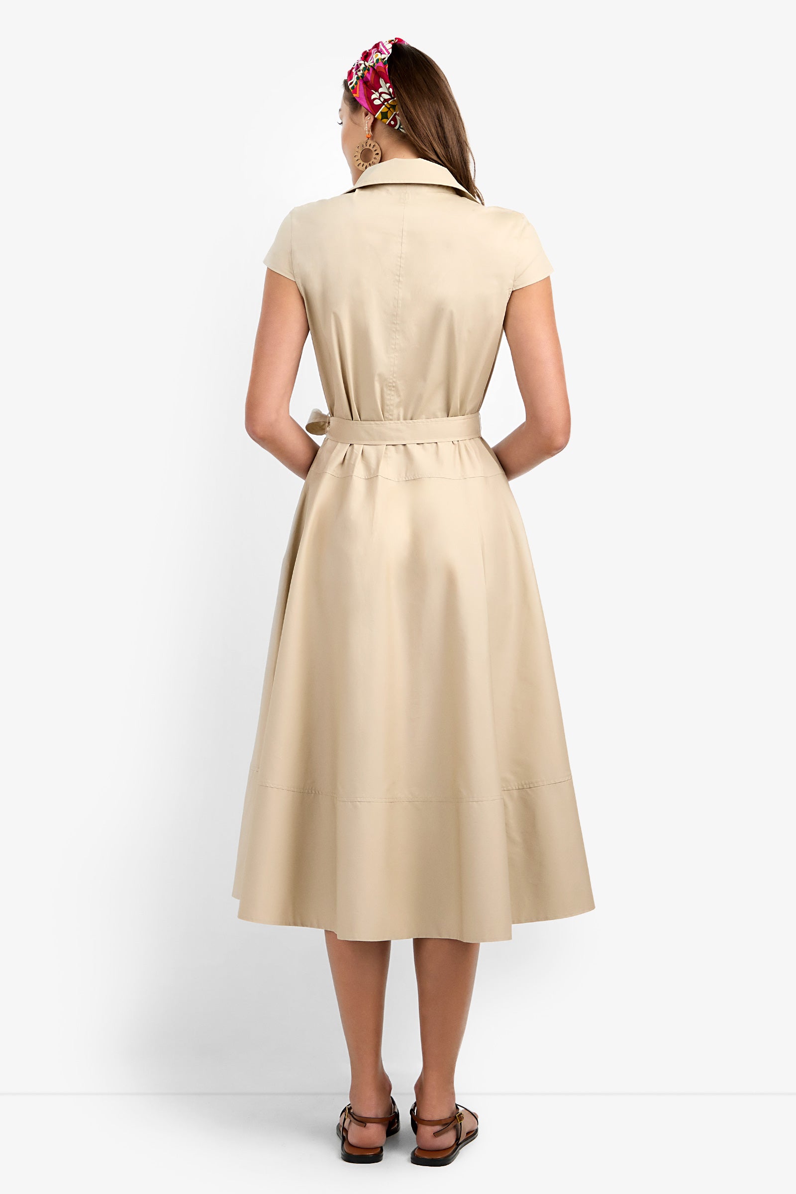 A woman faces away from the camera in a Delfina Dress—a short-sleeve beige belted midi—paired with brown sandals and a vibrant hair accessory, set against a plain white backdrop.