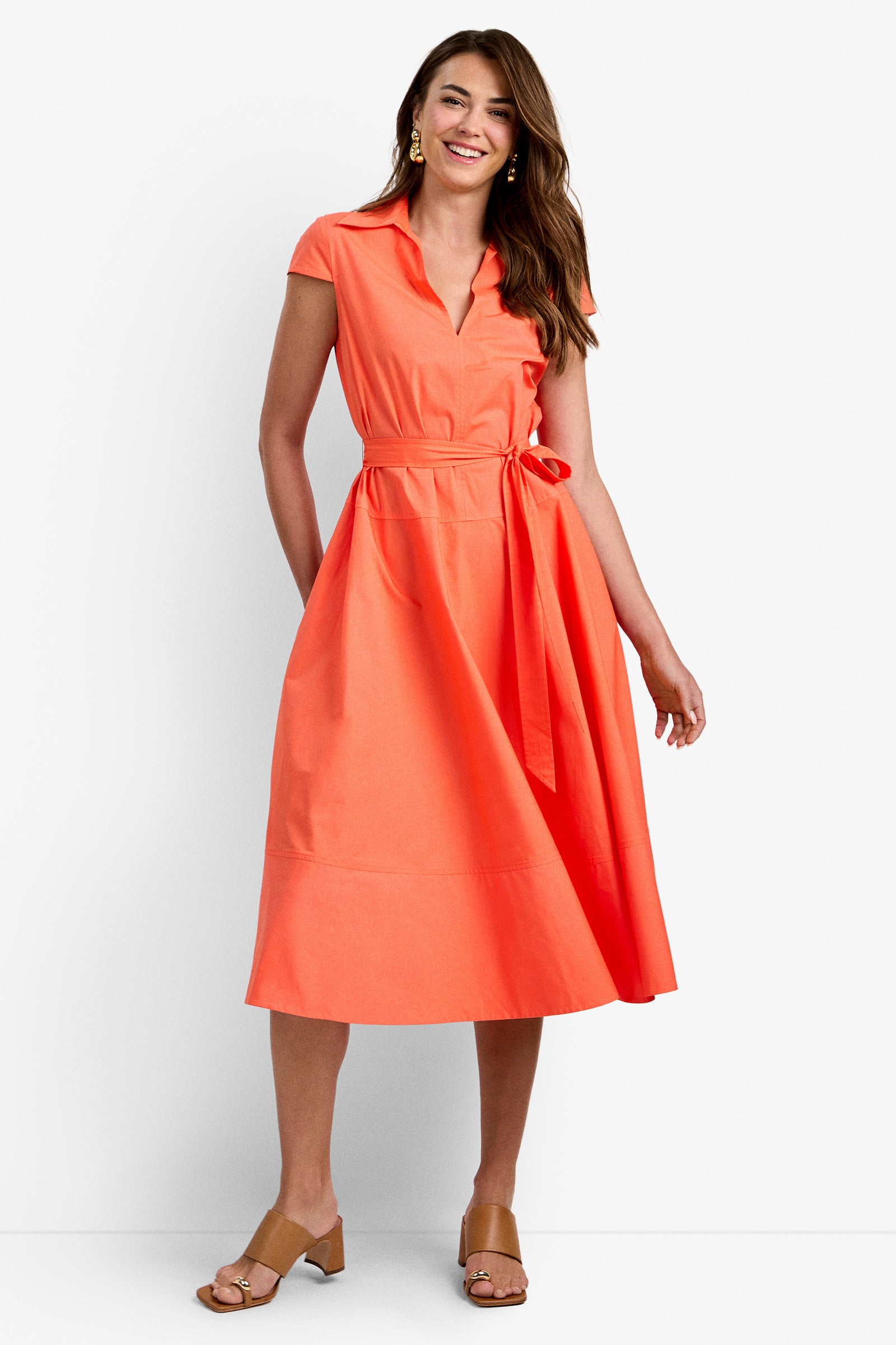 A woman with long brown hair smiles in the DELFINA DRESS—a coral, collared, short-sleeve midi with a waist tie and pockets—styled with tan heeled sandals and gold earrings.