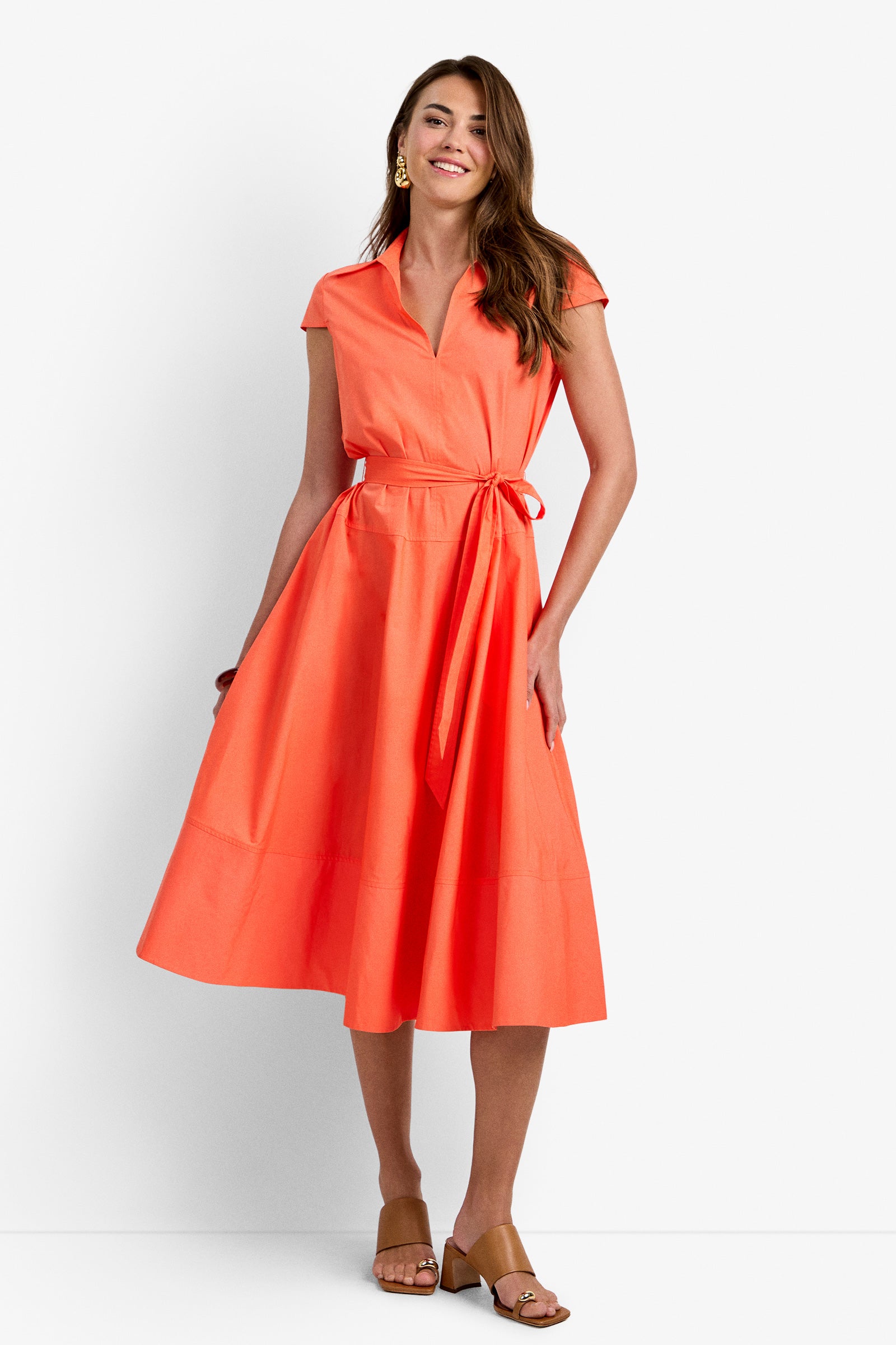 A woman in the DELFINA DRESS—a bright orange, short-sleeve, collared midi wrap with a tie belt—smiles while wearing tan heeled sandals against a plain white background.