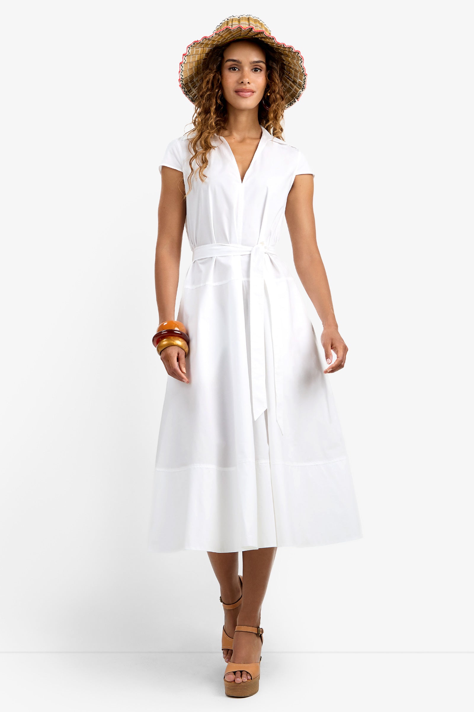 A woman in the white, short-sleeved, belted Delfina Dress, brown platform sandals, straw hat, and bracelets stands smiling against a plain white background.