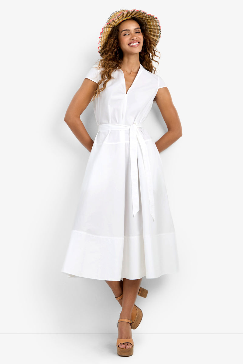 A woman smiles and poses with crossed feet in the Delfina Dress—a white, short-sleeve, belted midi dress—paired with tan platform sandals and a wide-brimmed plaid sun hat with multicolored trim. White background.
