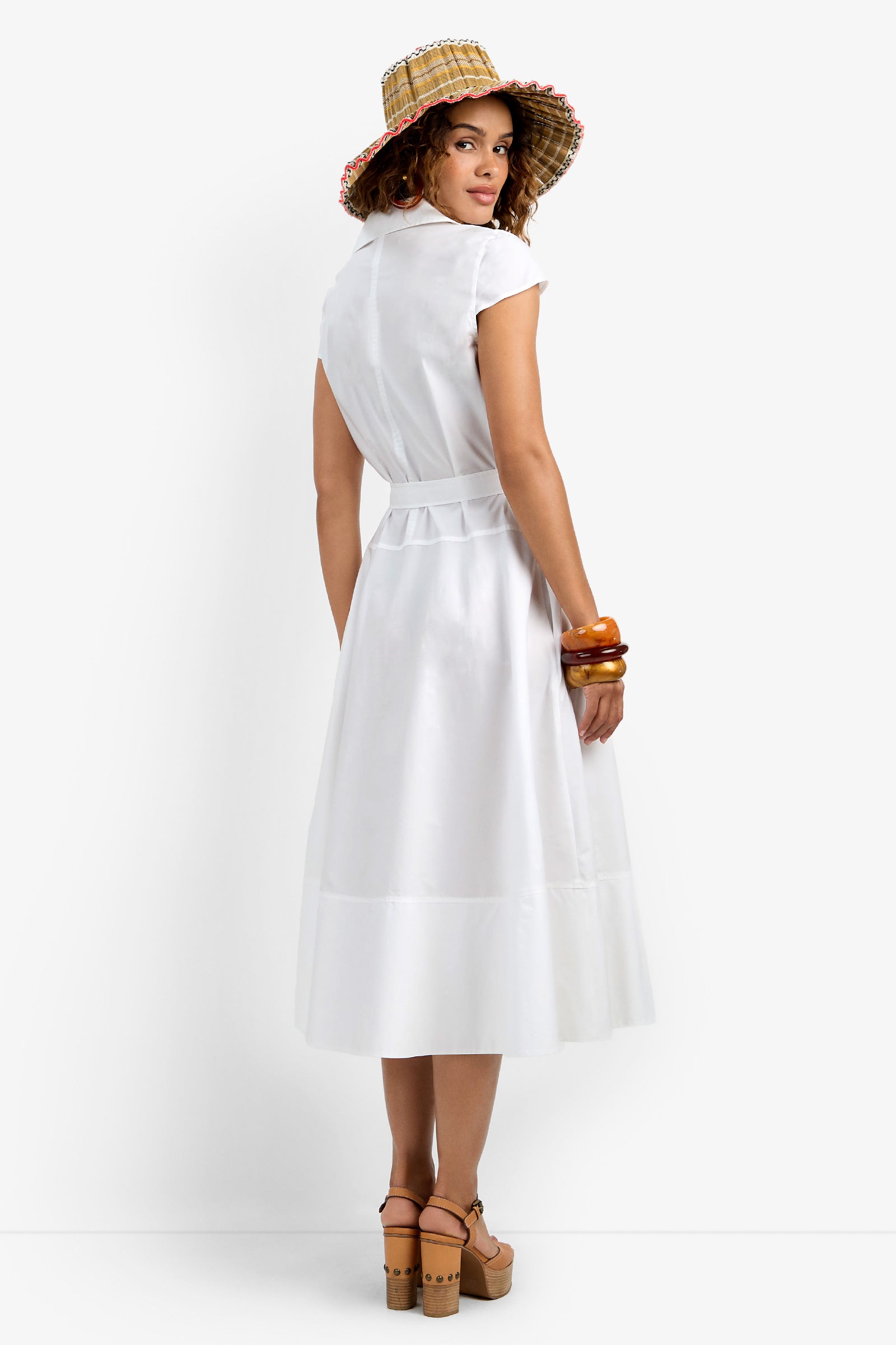 A woman in the Delfina Dress—a white, belted, short-sleeve midi—paired with a straw hat with a colorful brim, brown platform sandals, and chunky bracelets stands facing away, looking over her shoulder against a white background.