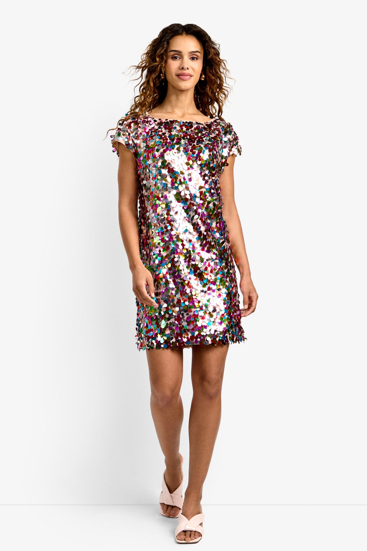 A woman with wavy hair wears the Oceana Dress in MULTI, a short-sleeved, knee-length style adorned with large colorful sequins, paired with light pink open-toe heels against a white background.