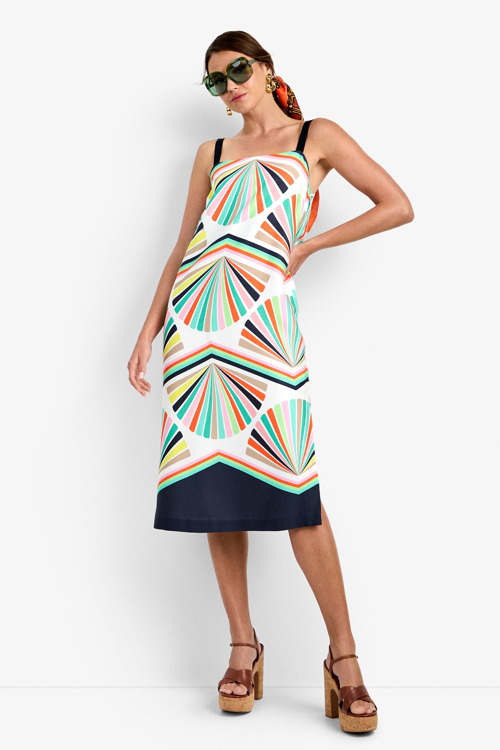 A woman wearing sunglasses, the Bayside Dress—a colorful geometric-patterned sleeveless dress with thin straps—tan platform sandals, and a patterned hair scarf poses with one hand on her hip against a white background.