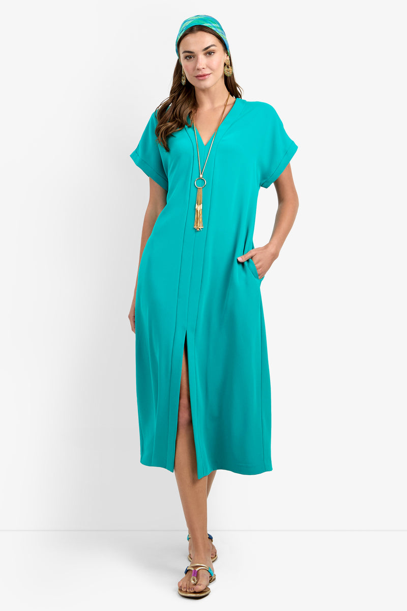 A woman wearing the OASIS DRESS—a turquoise midi with short sleeves, front slit, and pockets—paired with a matching headband, long pendant necklace, and sandals, stands against a white background.