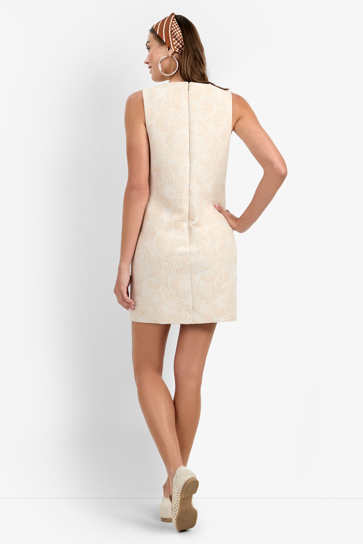 Woman facing away, wearing the SANDIE DRESS—a sleeveless beige mini dress with a subtle floral pattern—paired with wedge sandals, large hoop earrings, and a brown patterned headband. She stands against a plain white background.