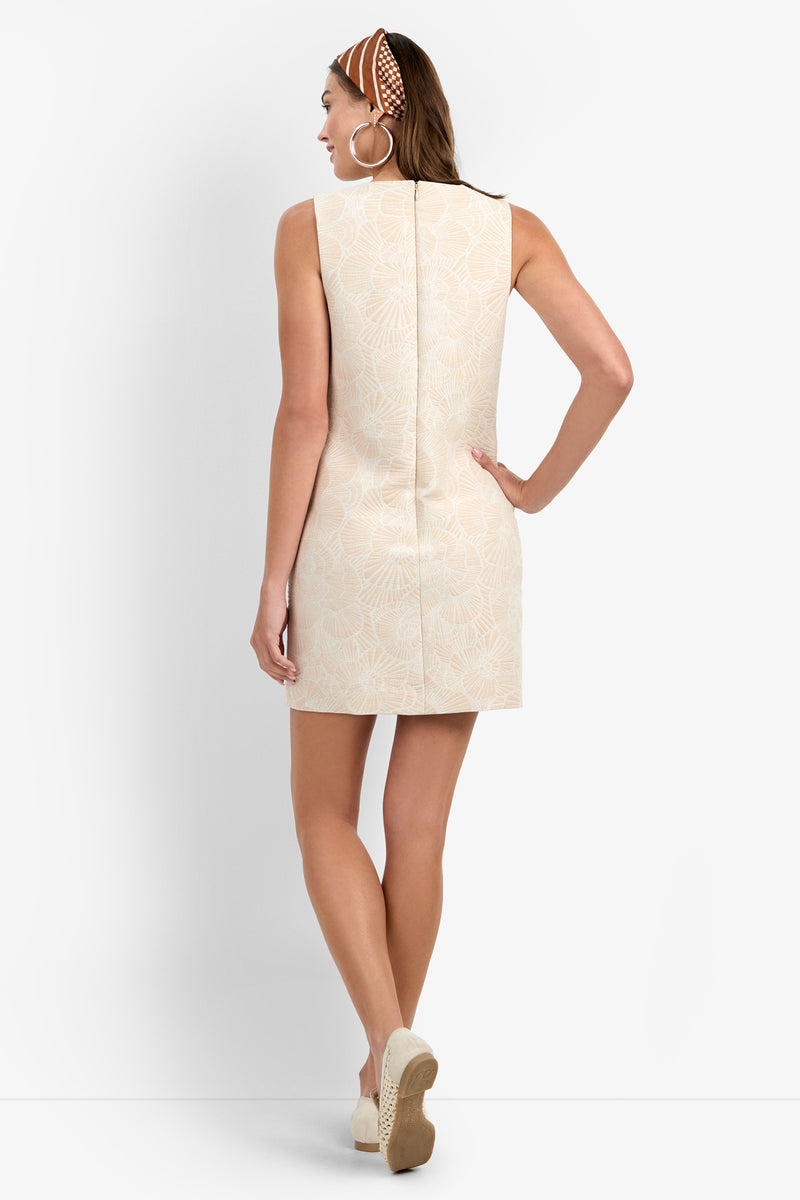 Woman facing away, wearing the SANDIE DRESS—a sleeveless beige mini dress with a subtle floral pattern—paired with wedge sandals, large hoop earrings, and a brown patterned headband. She stands against a plain white background.