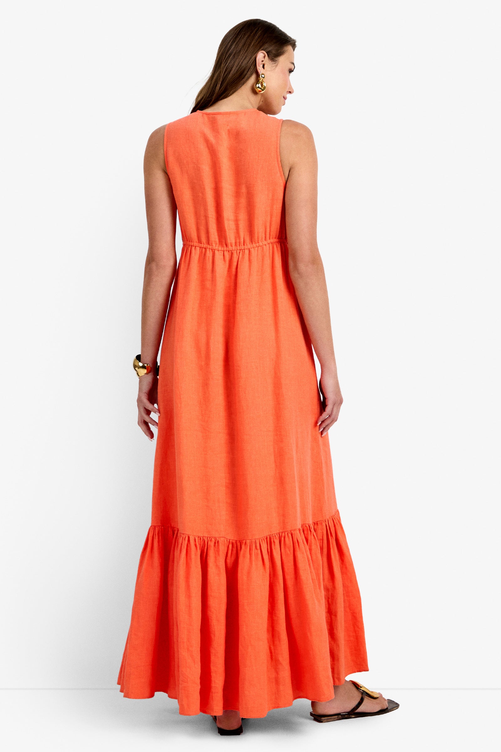A woman faces away, wearing the Lobster Dress—a sleeveless, ankle-length bright orange dress with a gathered waist and ruffled hem—paired with flat sandals, large gold hoop earrings, and a gold bracelet.