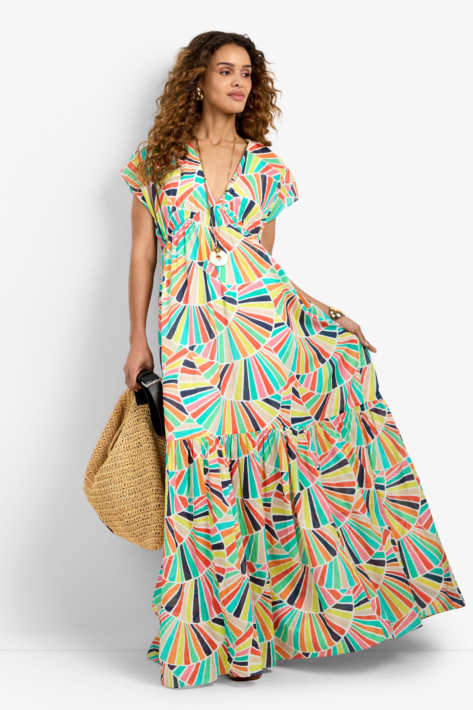 A woman with curly hair wears the CAPECOD DRESS in MULTI and holds a large woven straw bag. She stands confidently against a plain white background.