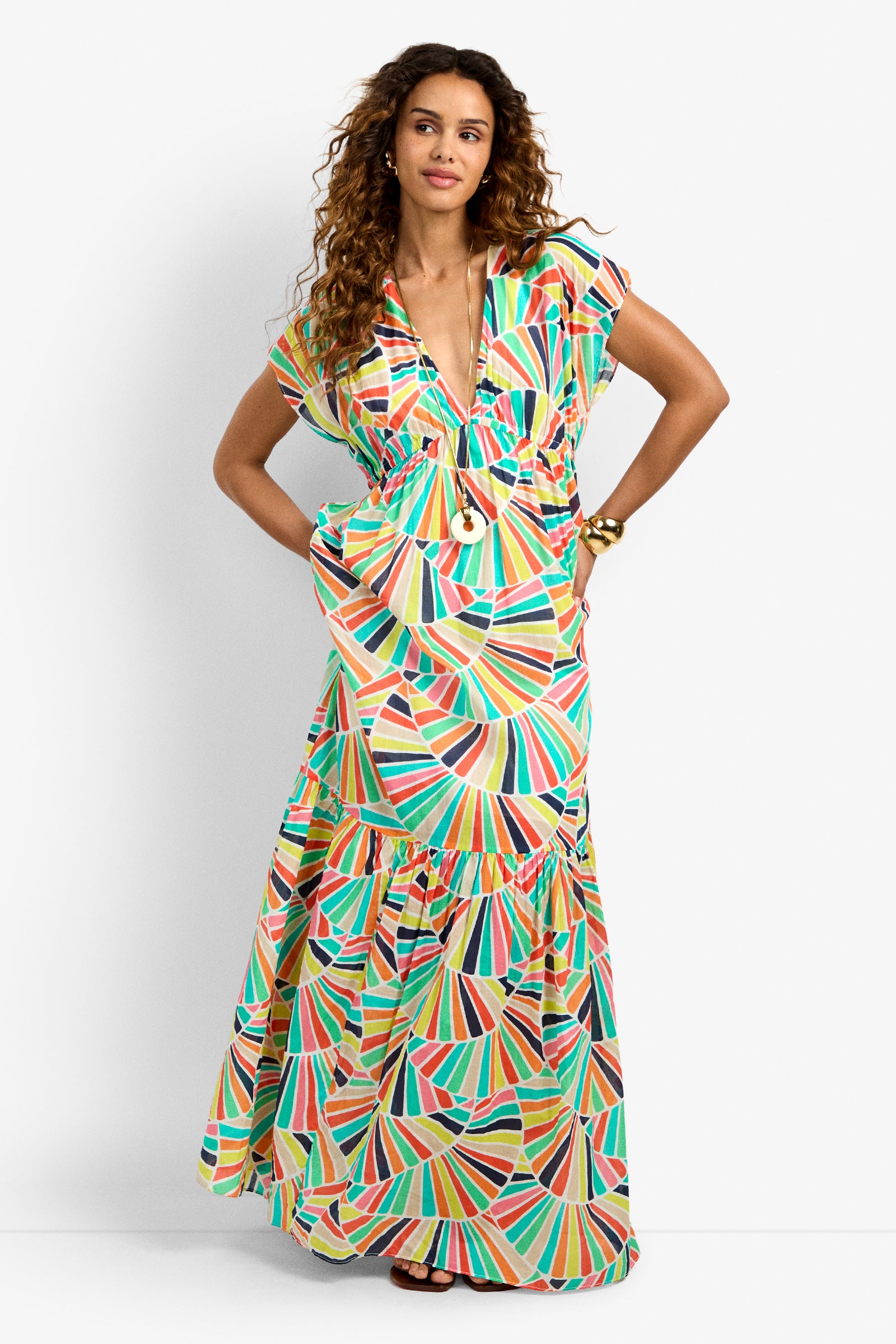 A woman with long curly hair wears the CAPECOD DRESS, a colorful geometric-patterned maxi with a deep V-neck and ruffled hem, accessorized with a pendant necklace and bracelets, standing against a white background.