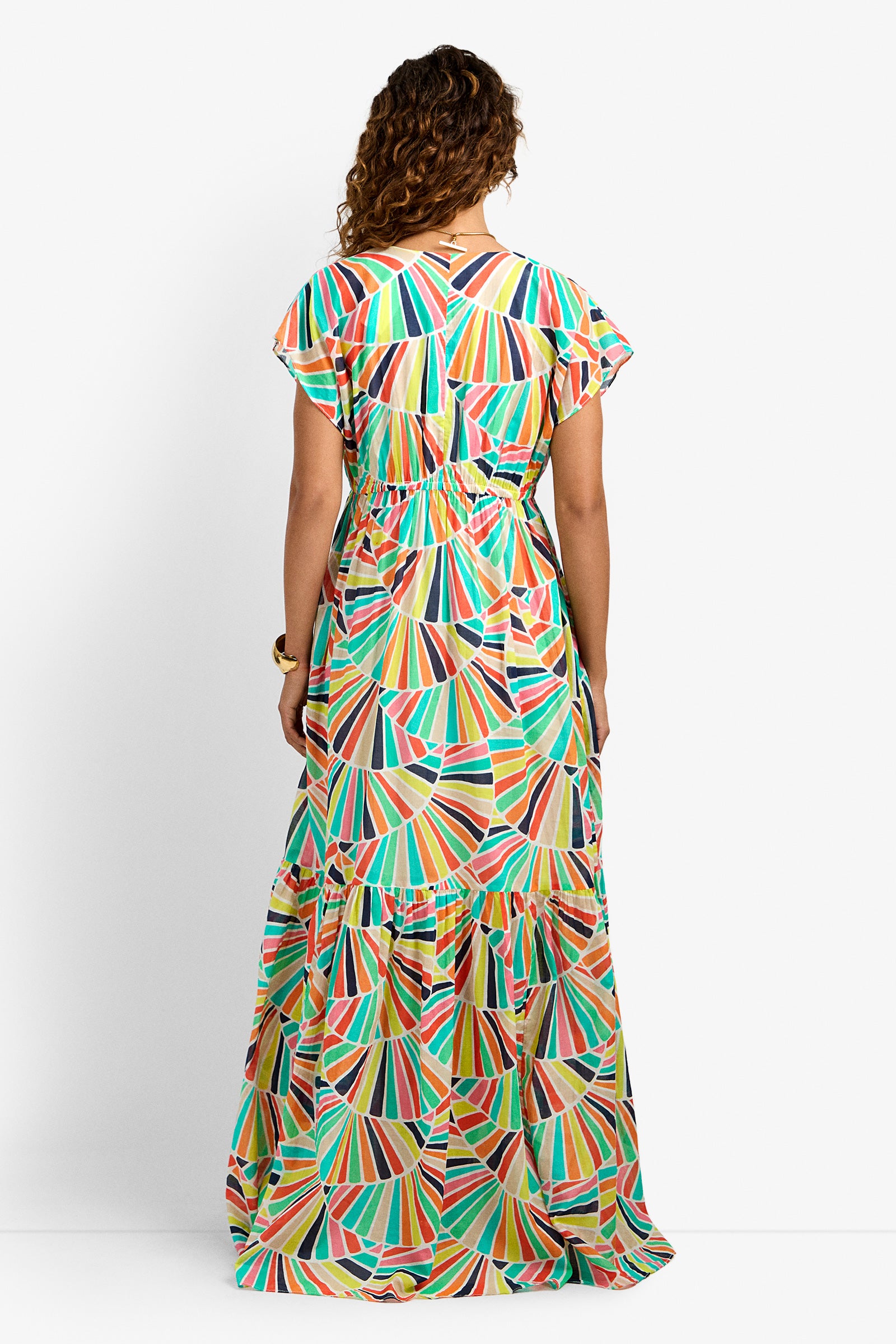 A person with curly hair stands facing away, wearing the CAPECOD DRESS—a colorful, patterned maxi dress with short sleeves and a tiered skirt—set against a plain white background.