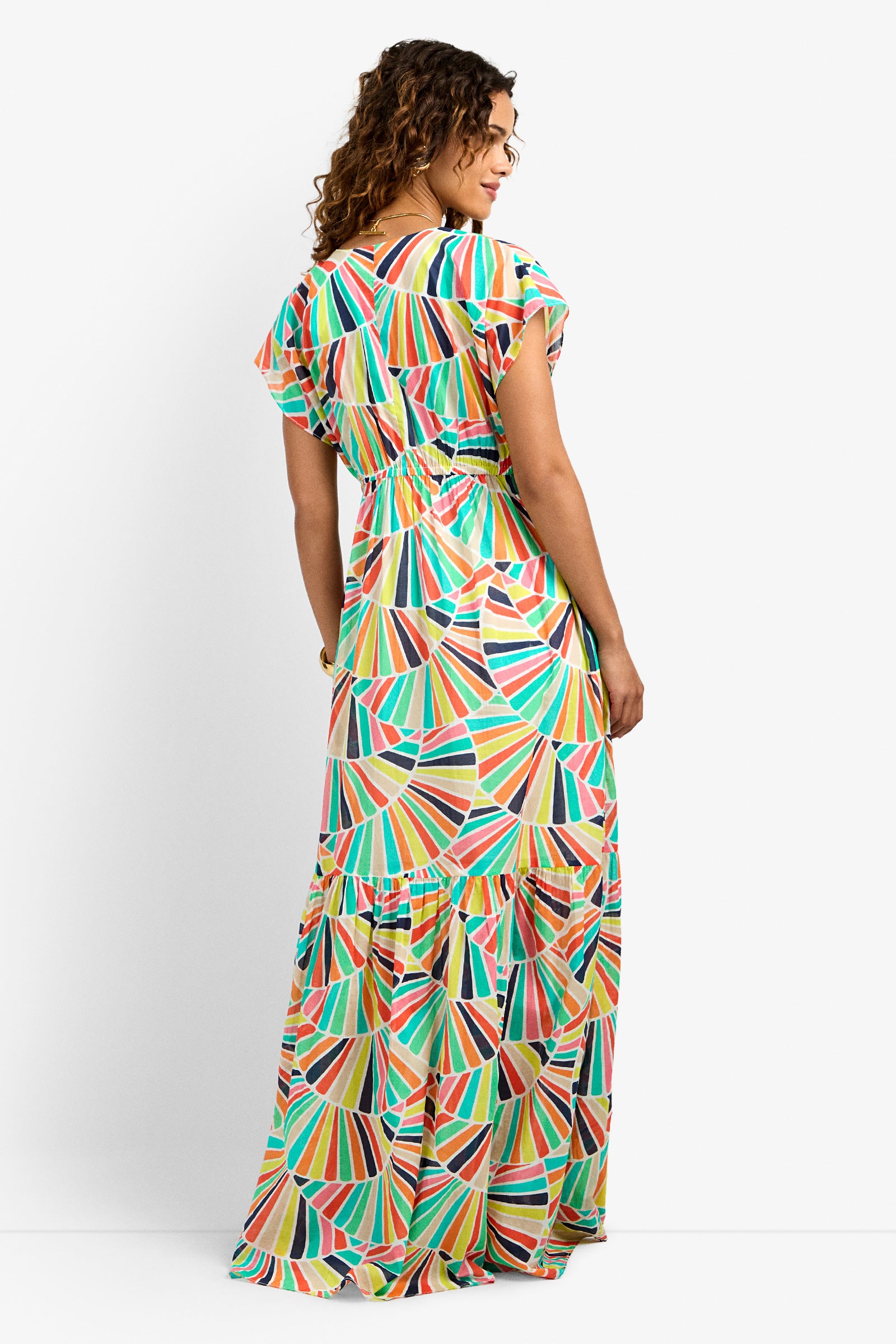 A woman with curly hair faces away, wearing the CAPECOD DRESS—a colorful, geometric-patterned maxi with short sleeves and a tiered skirt—against a plain white background.