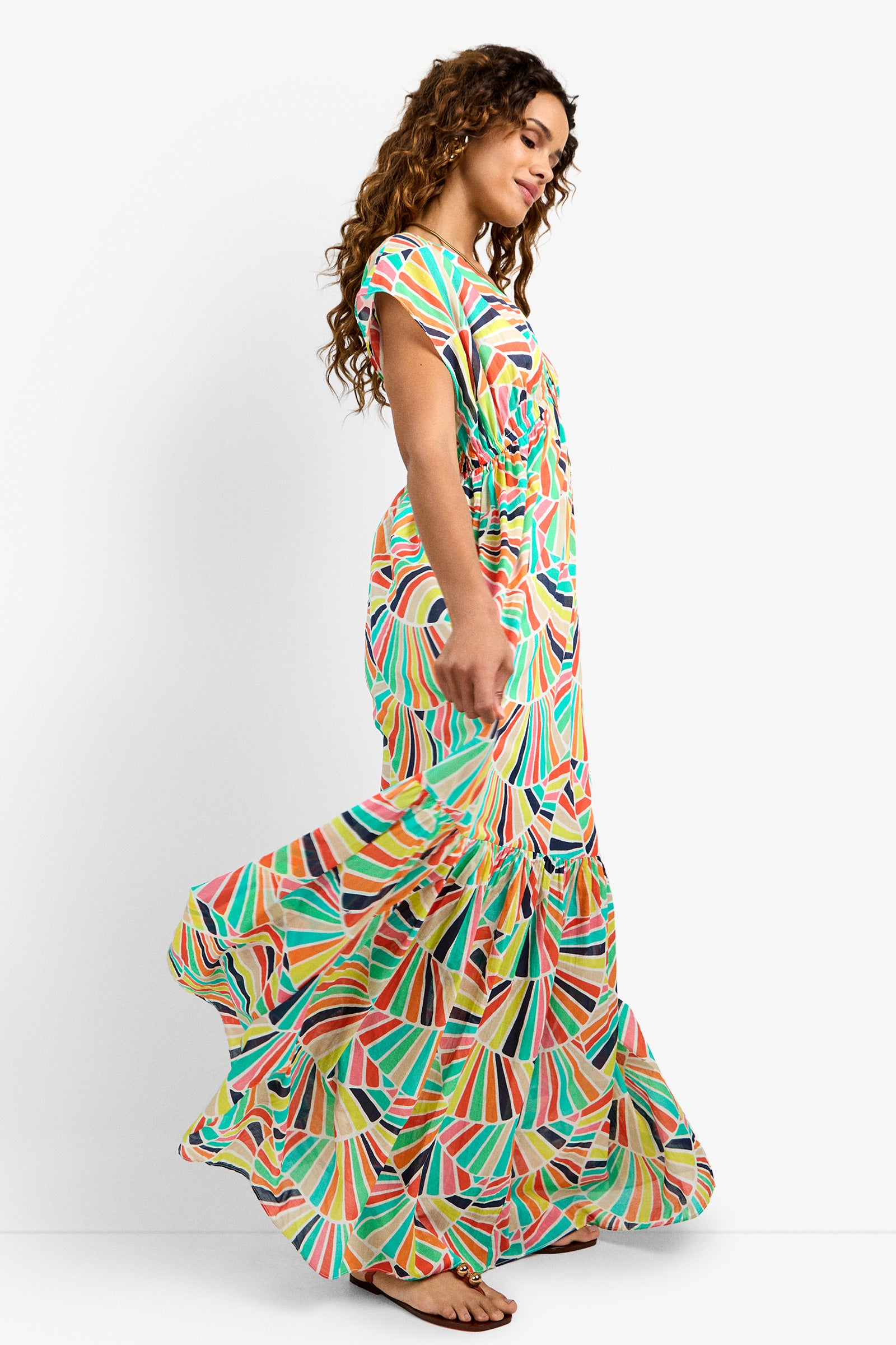 A woman with long curly hair wears the CAPECOD DRESS, a colorful geometric-patterned maxi, and sandals as she walks to the right. The flowing dress stands out against a plain white background.