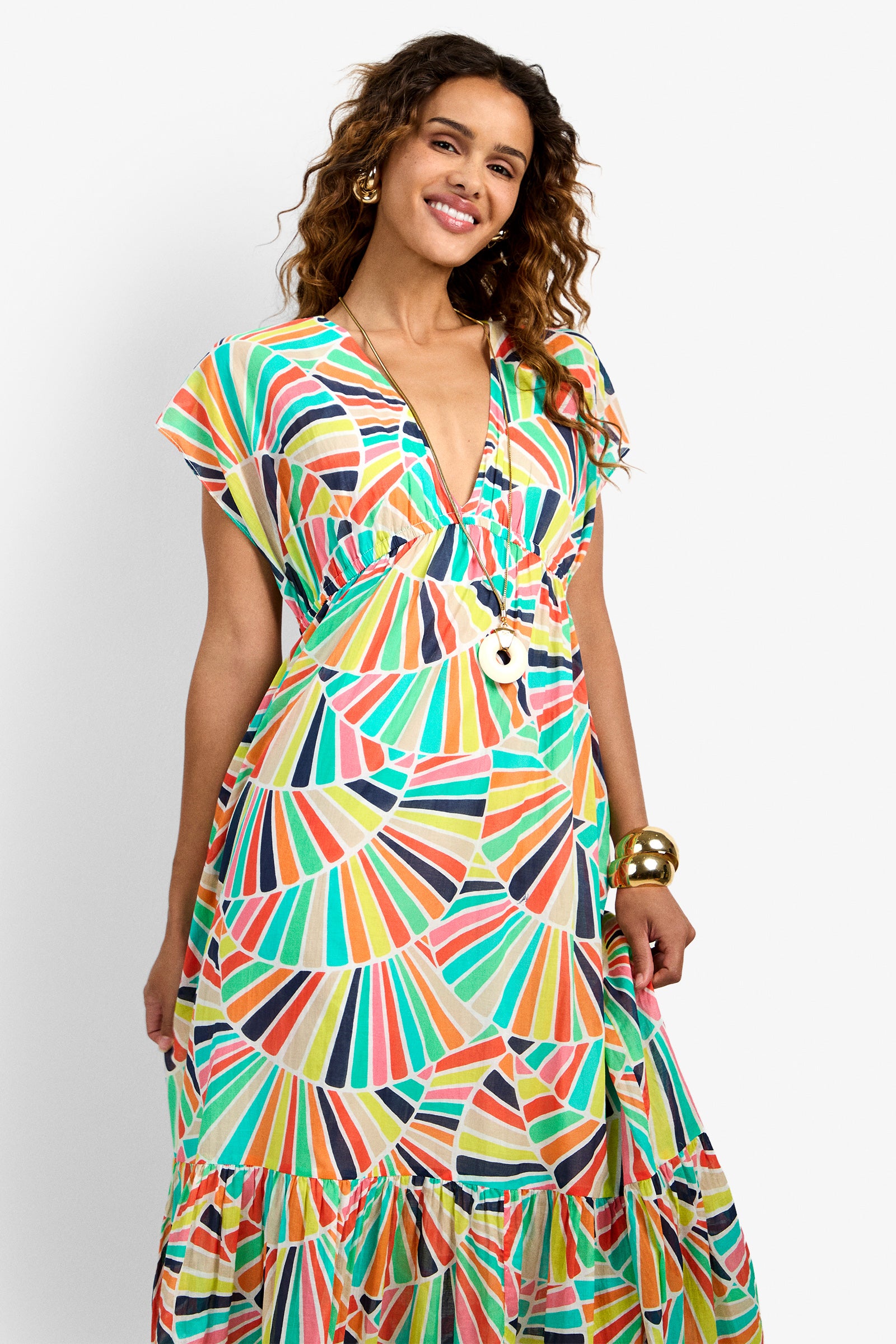 A woman smiles at the camera wearing the CAPECOD DRESS, a colorful patterned maxi with a V-neckline. She has curly hair, gold earrings, and a chunky bracelet, standing against a plain white background.