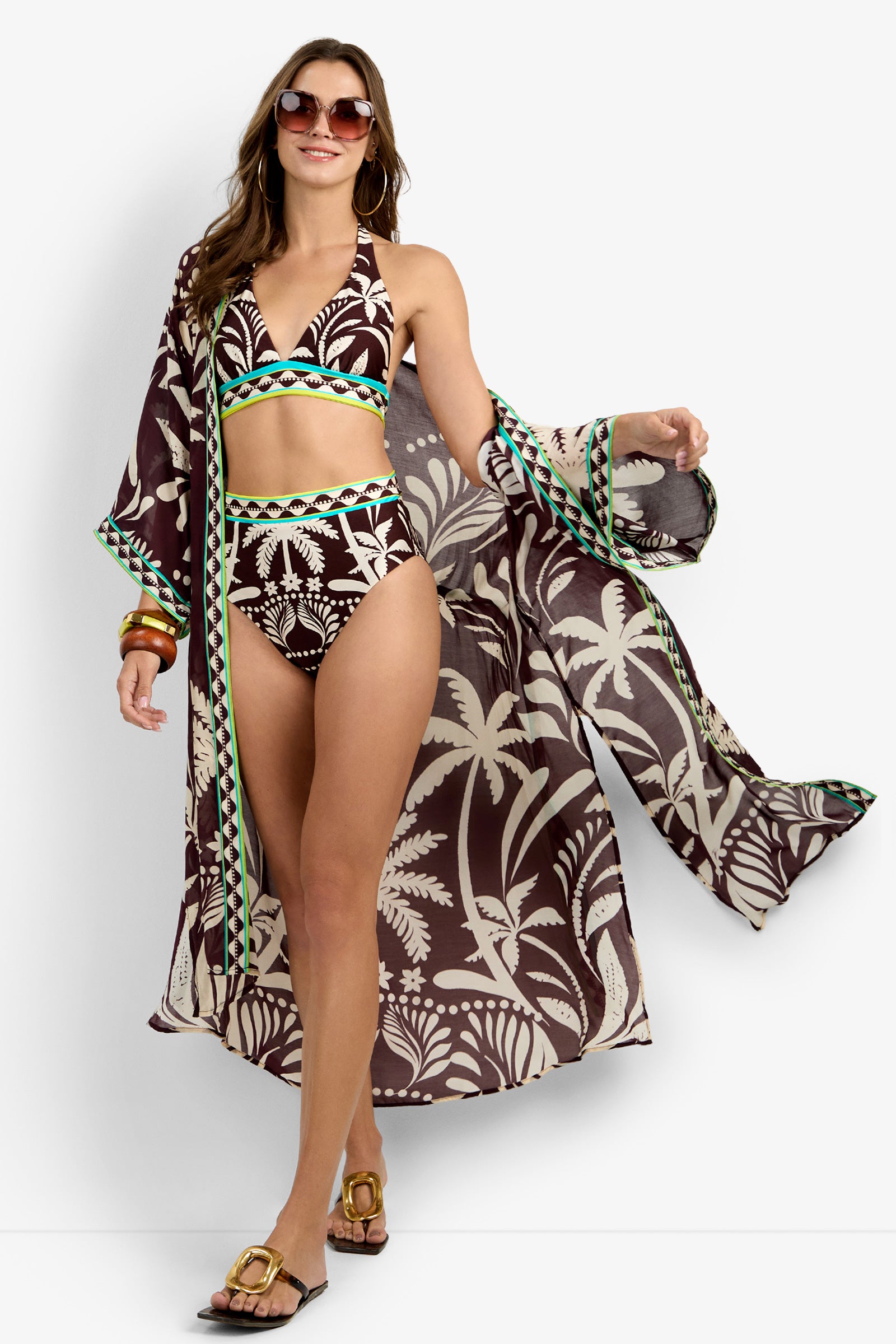 Woman wearing sunglasses, gold hoop earrings, bracelets, sandals, a brown and cream tropical print bikini, and the Kimono Maxi Duster cover-up with green trim. She confidently poses and smiles.