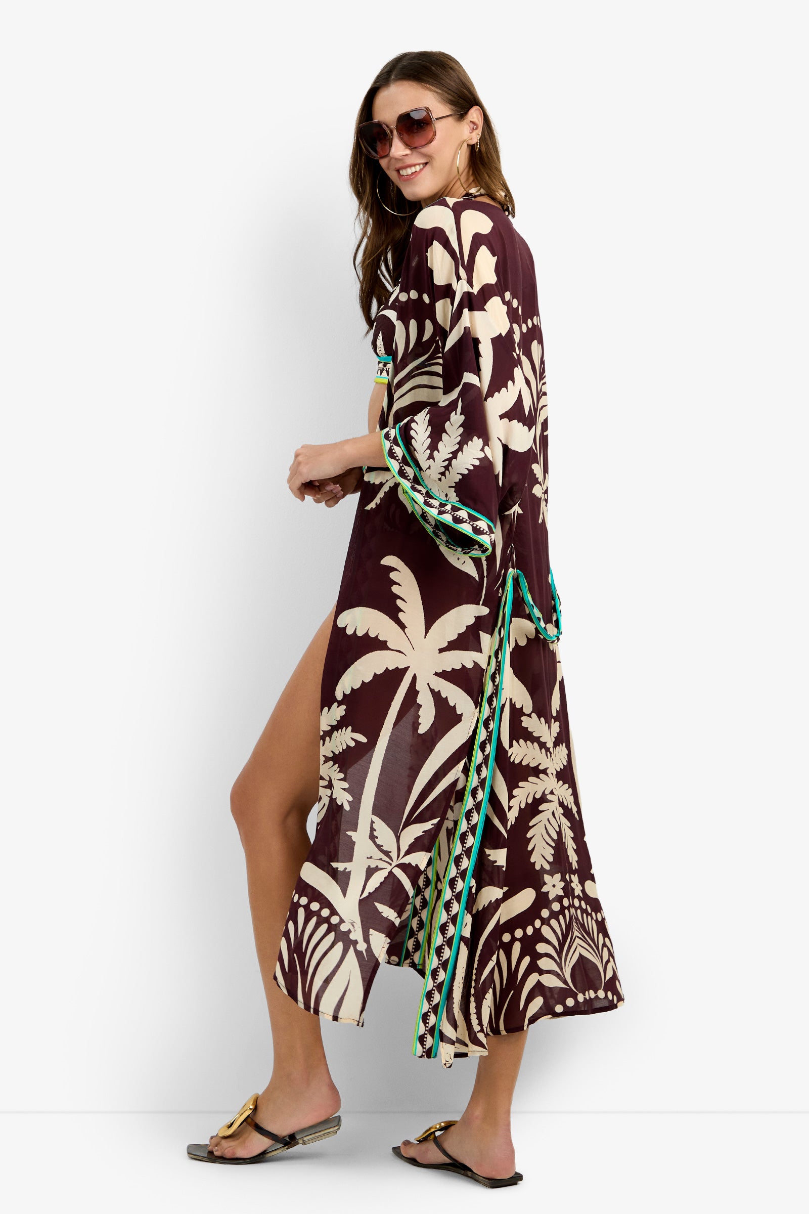 A woman poses side-on and smiles, wearing sunglasses, sandals, and the Kimono Maxi Duster—a long maroon beach cover-up with cream tropical patterns and green accents—against a plain white background.