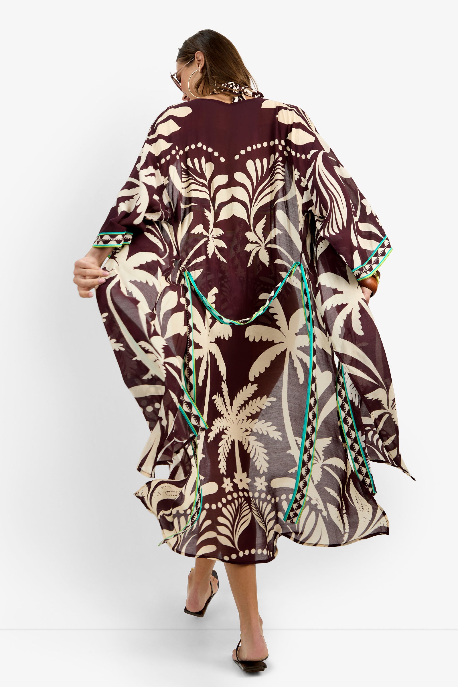 A woman with long hair, sunglasses, and sandals models the Kimono Maxi Duster, displaying its maroon and beige tropical print with green accents and sheer fabric as she holds out the sides to show off the pattern.