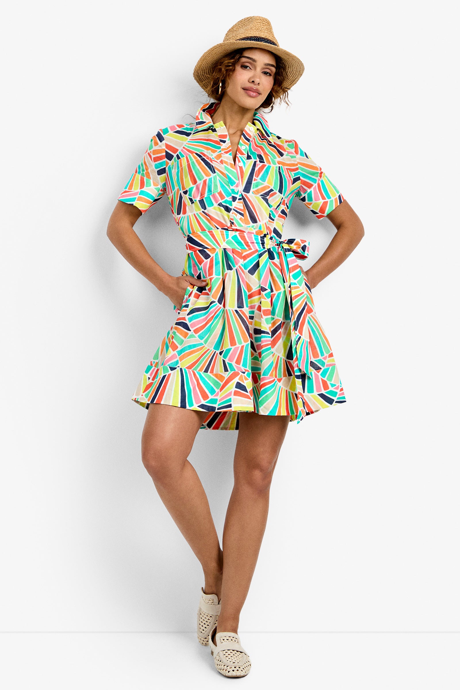 A woman models the Misola Dress in Multi, styled with a straw hat and woven slip-on shoes, standing against a white background with one hand on her hip and the other on her skirt.