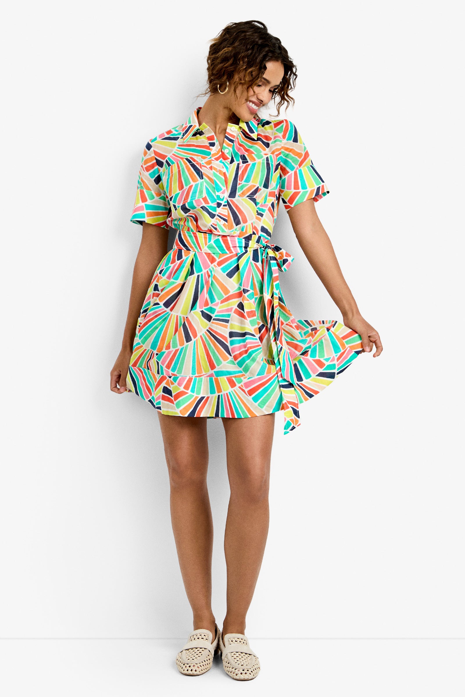Smiling woman wears the Misola Dress—a colorful, short-sleeve, knee-length dress with a geometric fan pattern and belted waist—with cream slip-on shoes, standing and holding the edge of her dress.
