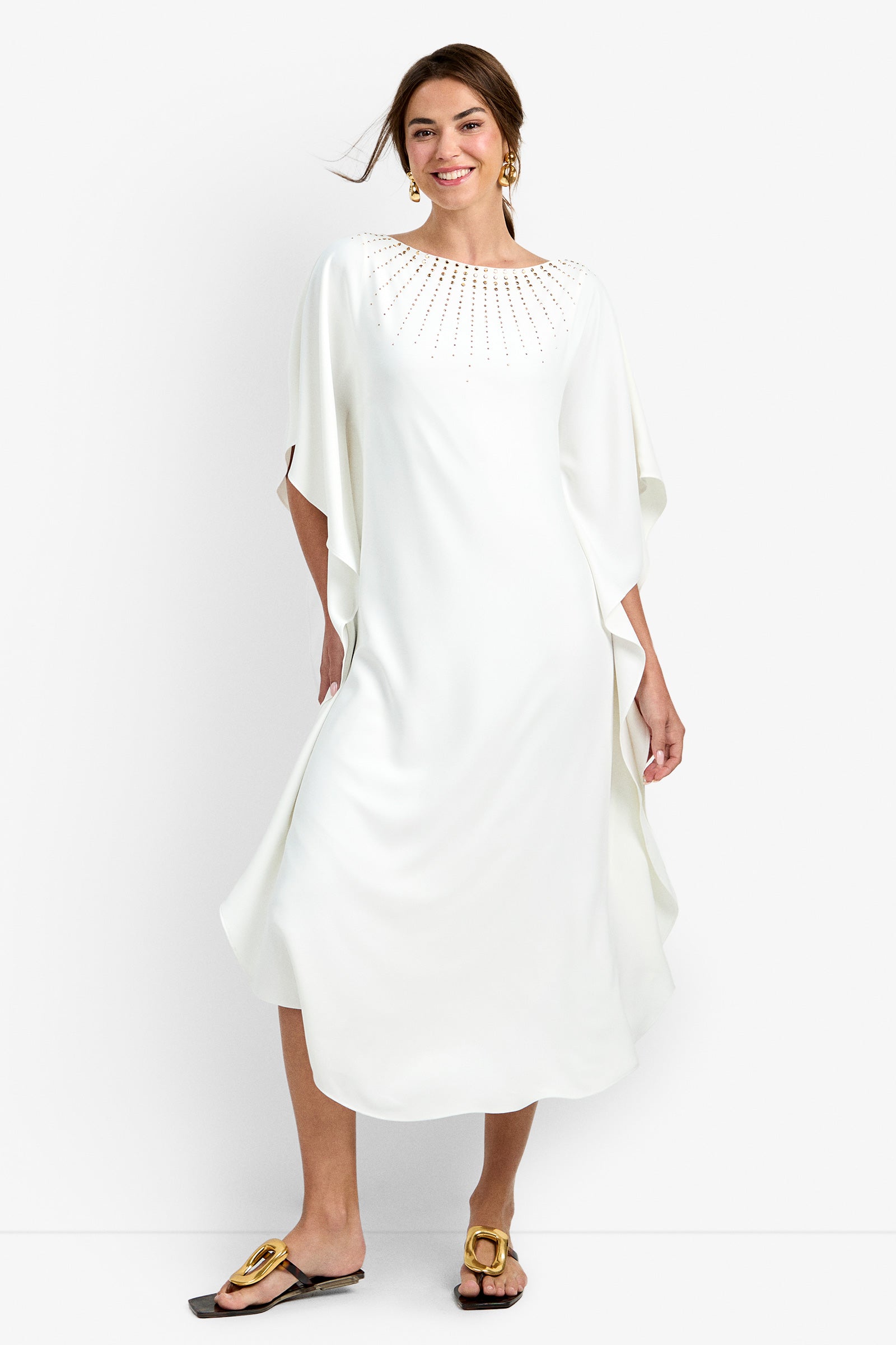 A woman smiles against a white background wearing the Neena Rhinestone Caftan Dress, featuring a flowy white midi silhouette with embellished neckline, styled with gold earrings and black flat sandals.
