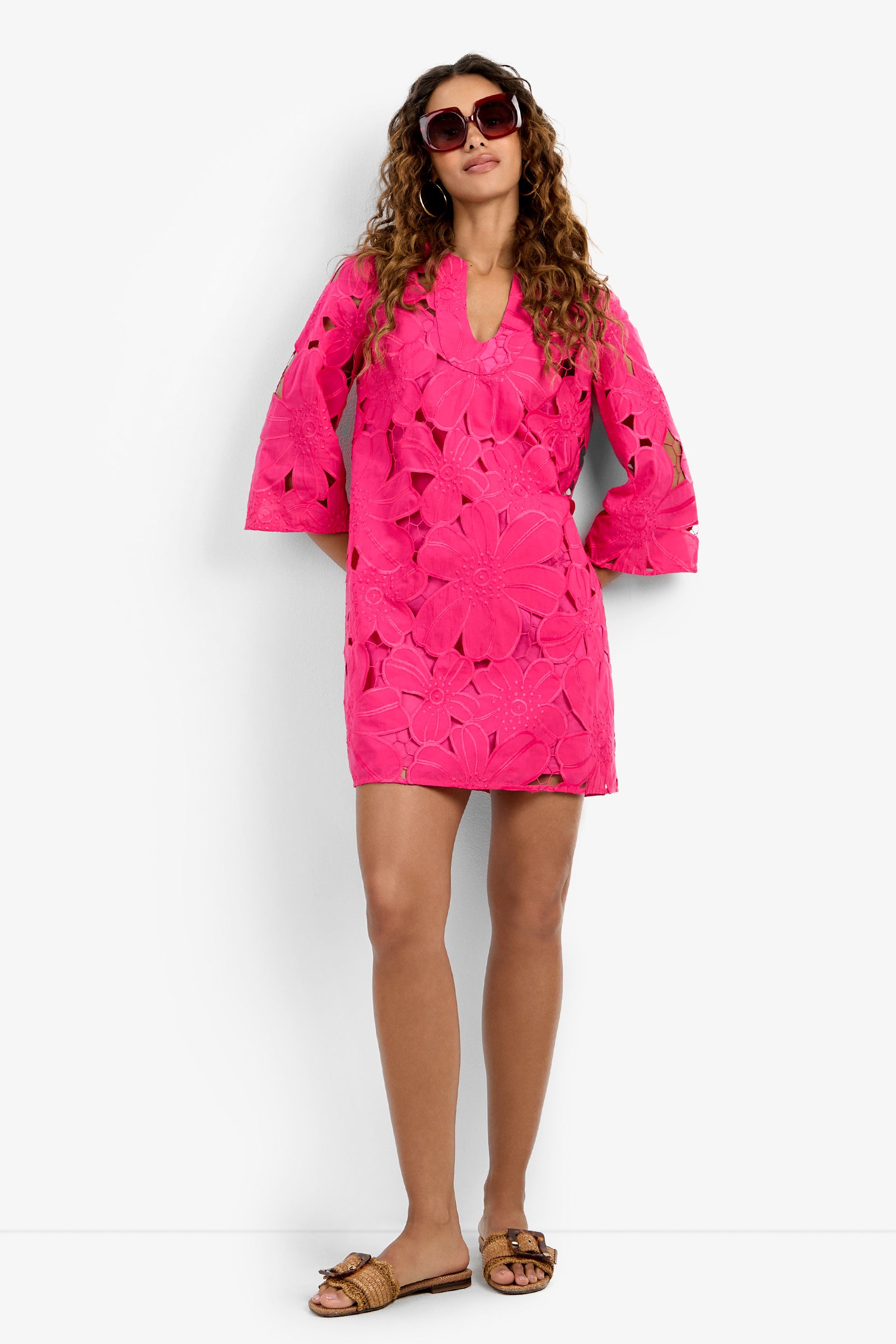 Woman with long curly hair wearing sunglasses, the Mangrove Dress—a bright pink floral-patterned dress with three-quarter sleeves—paired with brown flat sandals, standing against a plain white background.