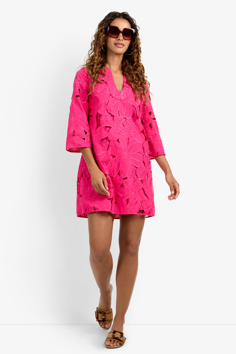 A woman in the Mangrove Dress—a bright pink, knee-length dress with floral cutout patterns—pairs it with brown sandals and sunglasses, walking confidently against a plain white background.