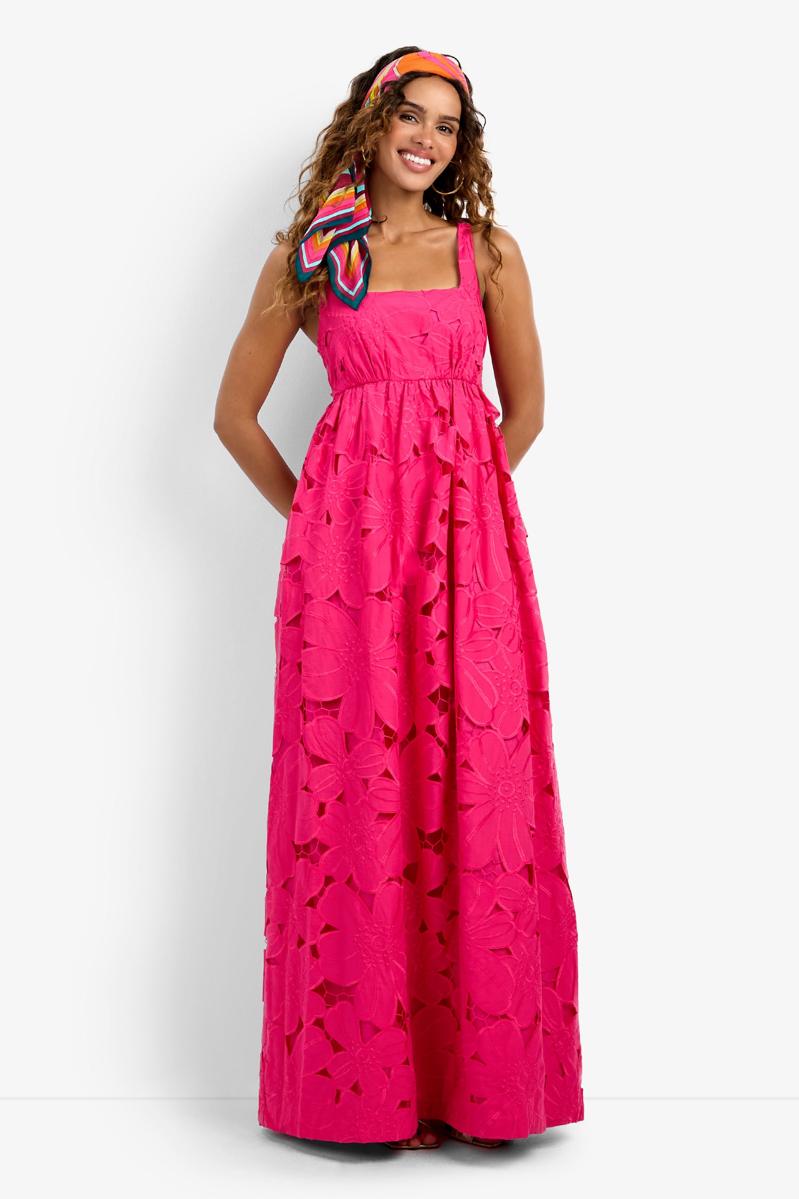 A woman stands smiling with her hands in the pockets of the Ariel Dress, a bright pink, sleeveless, floor-length dress featuring large floral patterns and eyelet details. She wears a colorful headscarf tied in her hair.