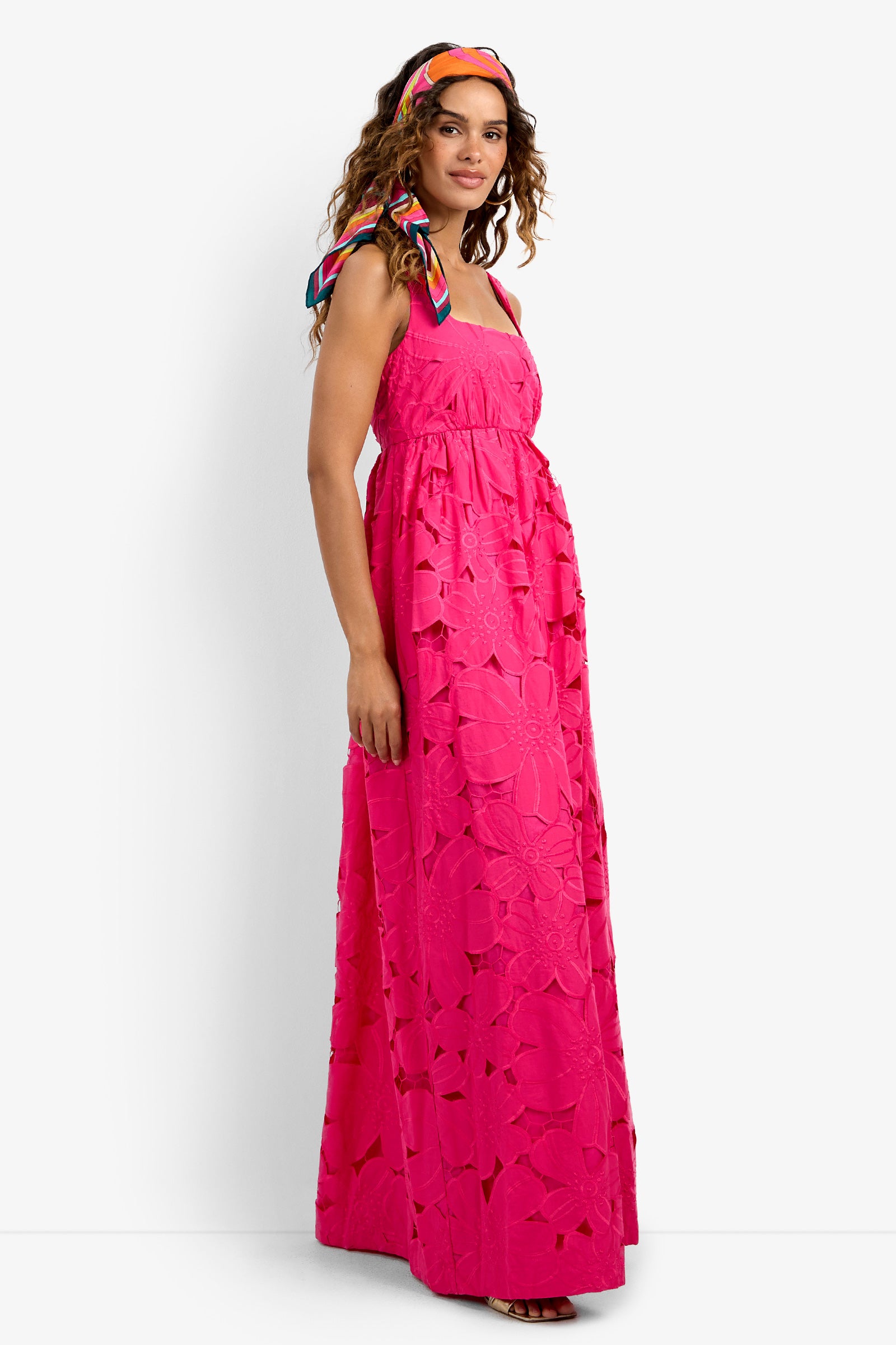Woman in the Ariel Dress—a bright pink, floral sleeveless maxi with spaghetti straps—stands against a white background. She smiles and looks to the side, her hair styled with a colorful scarf.