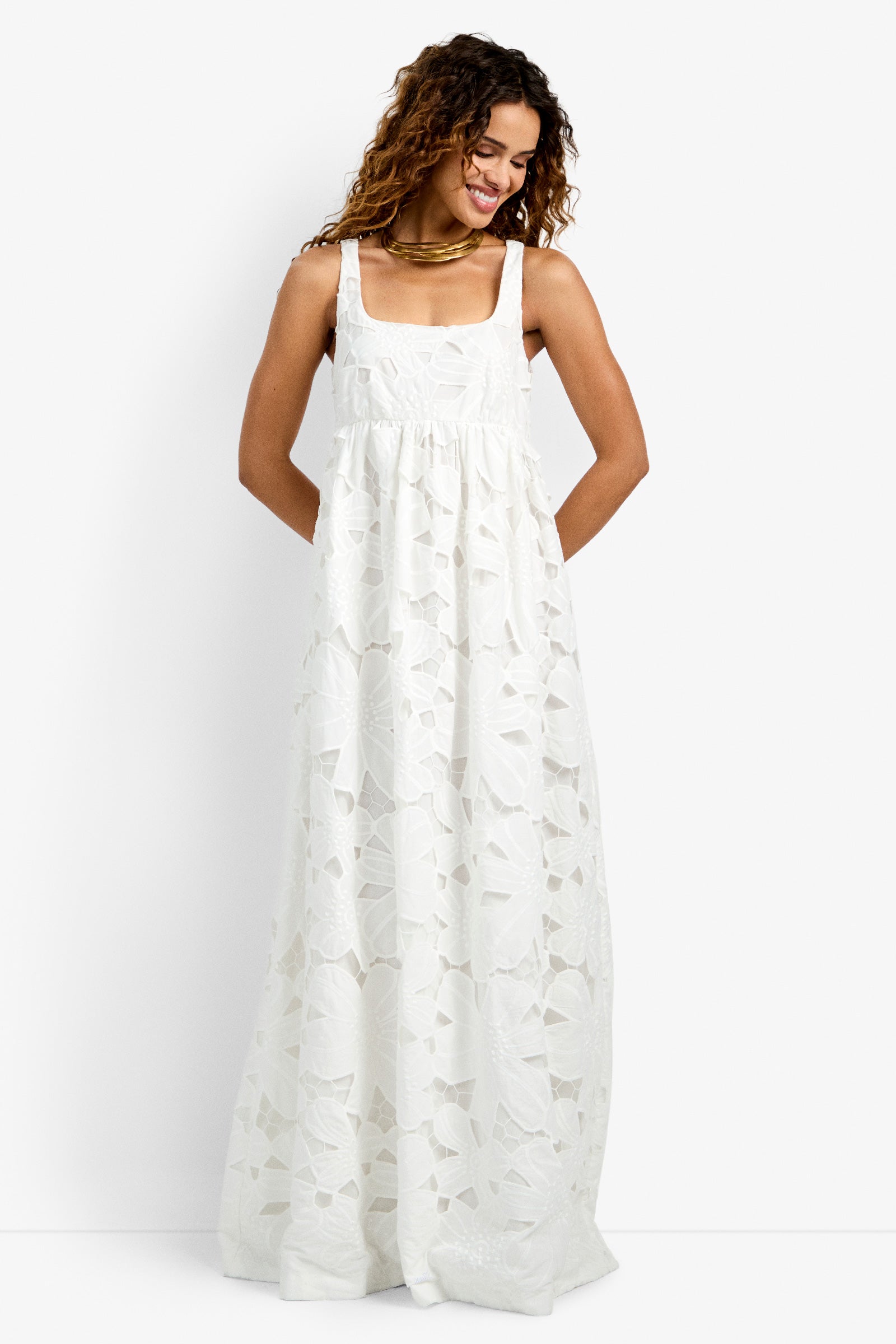 A woman with curly hair wears the Ariel Dress, a long, sleeveless white dress featuring a floral lace pattern. She smiles while looking down, standing against a plain white background.