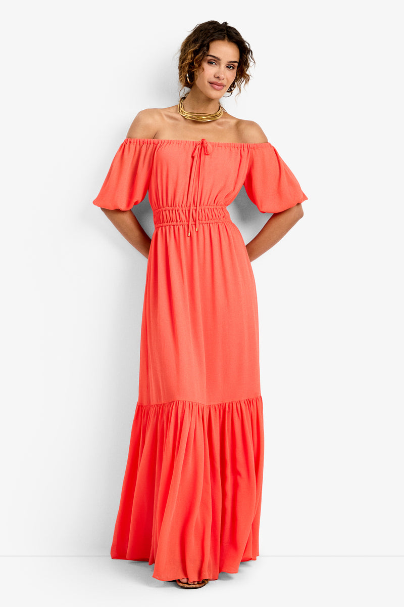 A woman with short curly hair wears the West Coast Dress—a coral, off-the-shoulder, short-sleeve, ankle-length dress featuring a gathered waist and ruffled hem—paired with gold jewelry, standing against a white background.