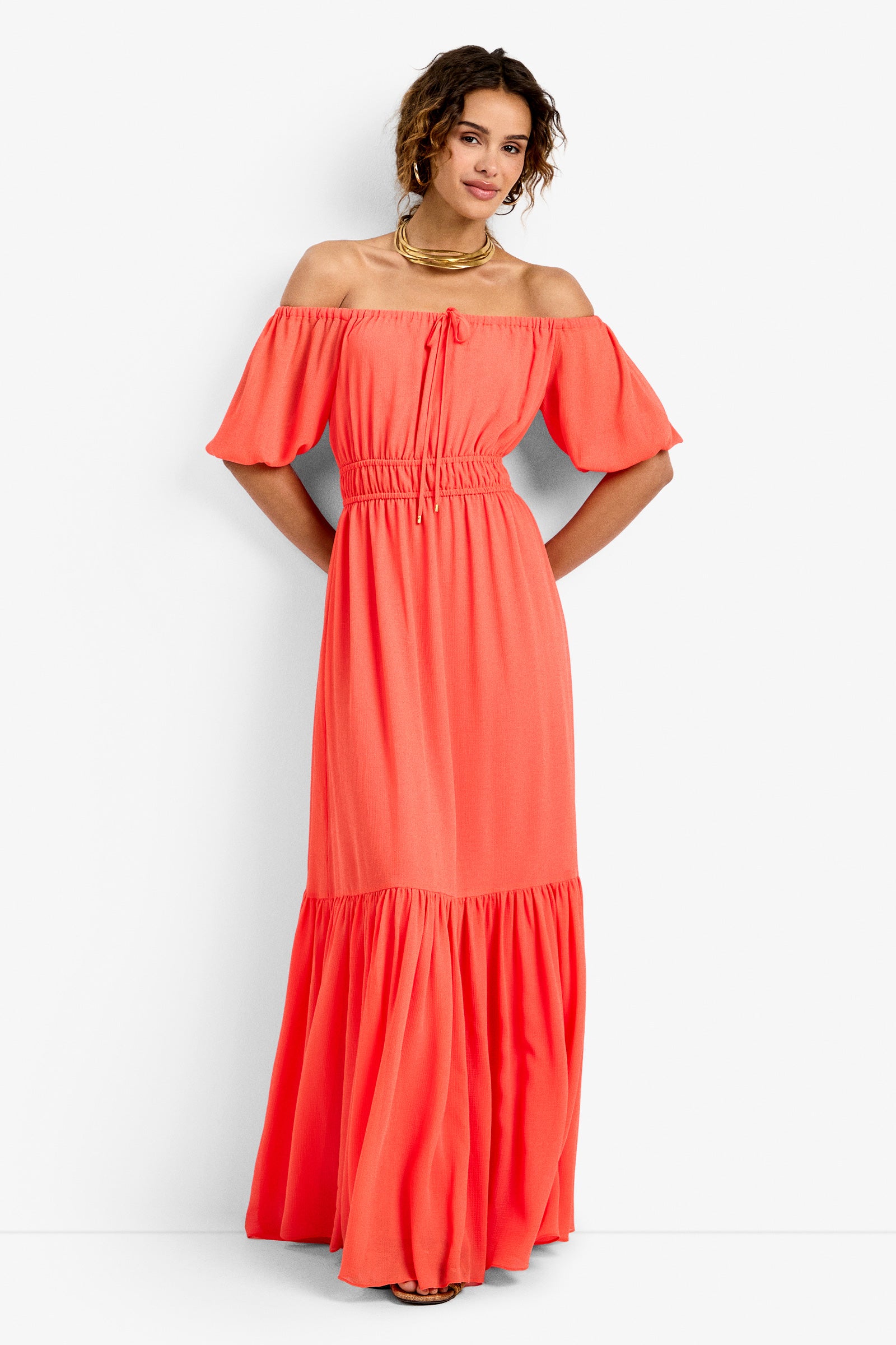 A woman with short curly hair wears the West Coast Dress—a coral, off-the-shoulder, short-sleeve, ankle-length dress featuring a gathered waist and ruffled hem—paired with gold jewelry, standing against a white background.