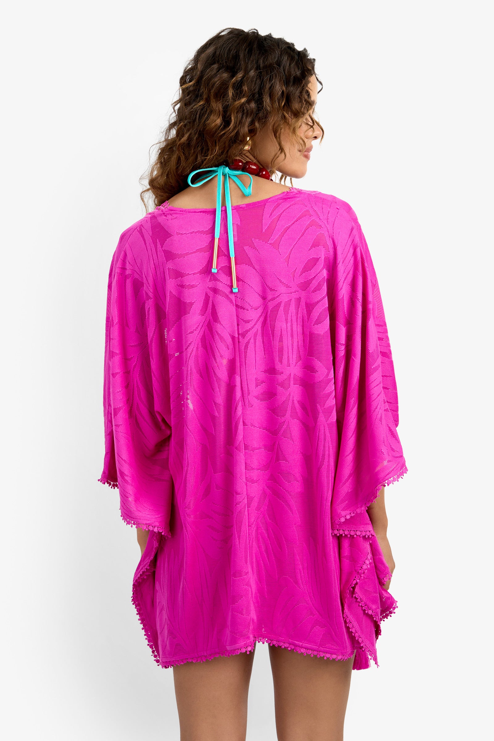 A woman with curly hair faces away, wearing a bright pink Kimono Cover-up featuring a tropical pattern, wide sleeves, and a scalloped hem. A blue swimsuit string is tied behind her neck.