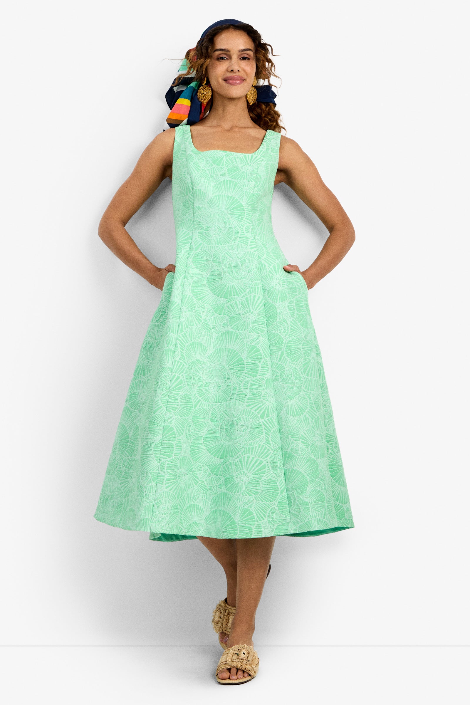 A woman smiles in the Valor 2 Dress in LUSH LAGOON, a sleeveless A-line midi with a subtle shell pattern. She has a colorful scarf in her curly hair, woven sandals, and her hands are tucked into the dress pockets.