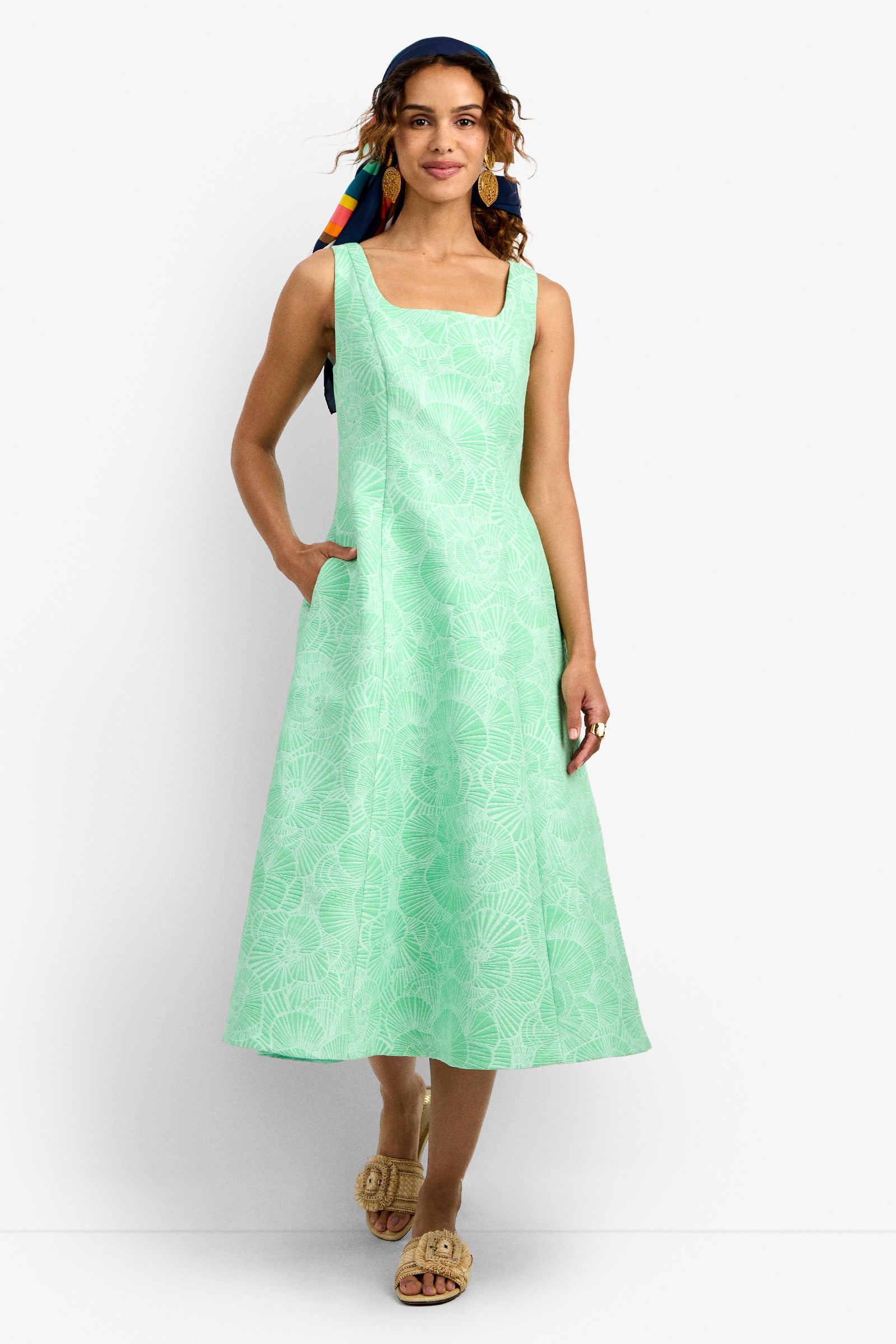 A woman smiles with one hand in her pocket, wearing the Valor 2 Dress—a light green, sleeveless A-line with a subtle leaf pattern—paired with tan sandals, gold earrings, and a colorful headscarf.