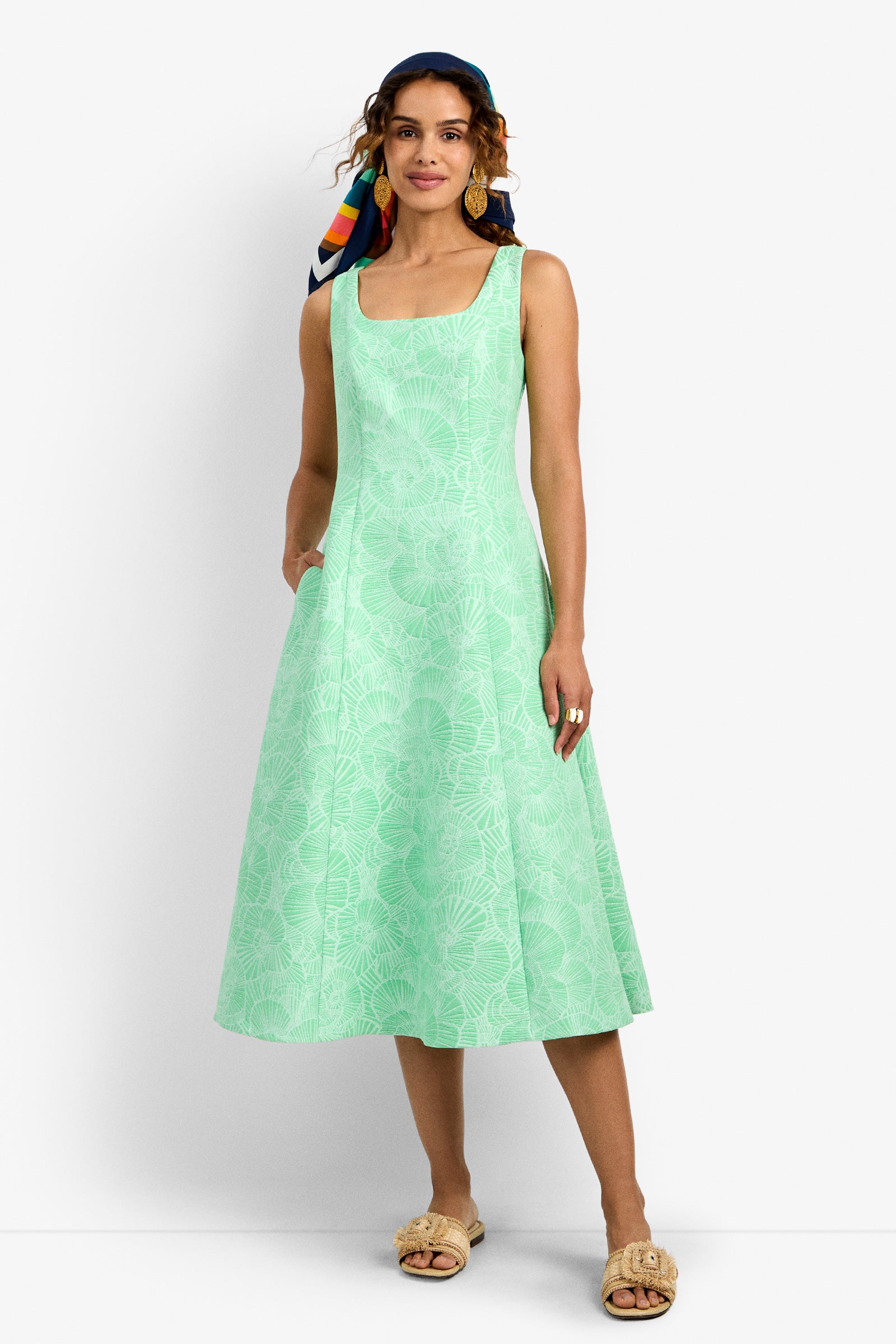 A woman stands against a plain white background wearing the Valor 2 Dress—a light green, sleeveless, knee-length dress with a subtle floral pattern—paired with tan sandals and a colorful scarf tied around her head.