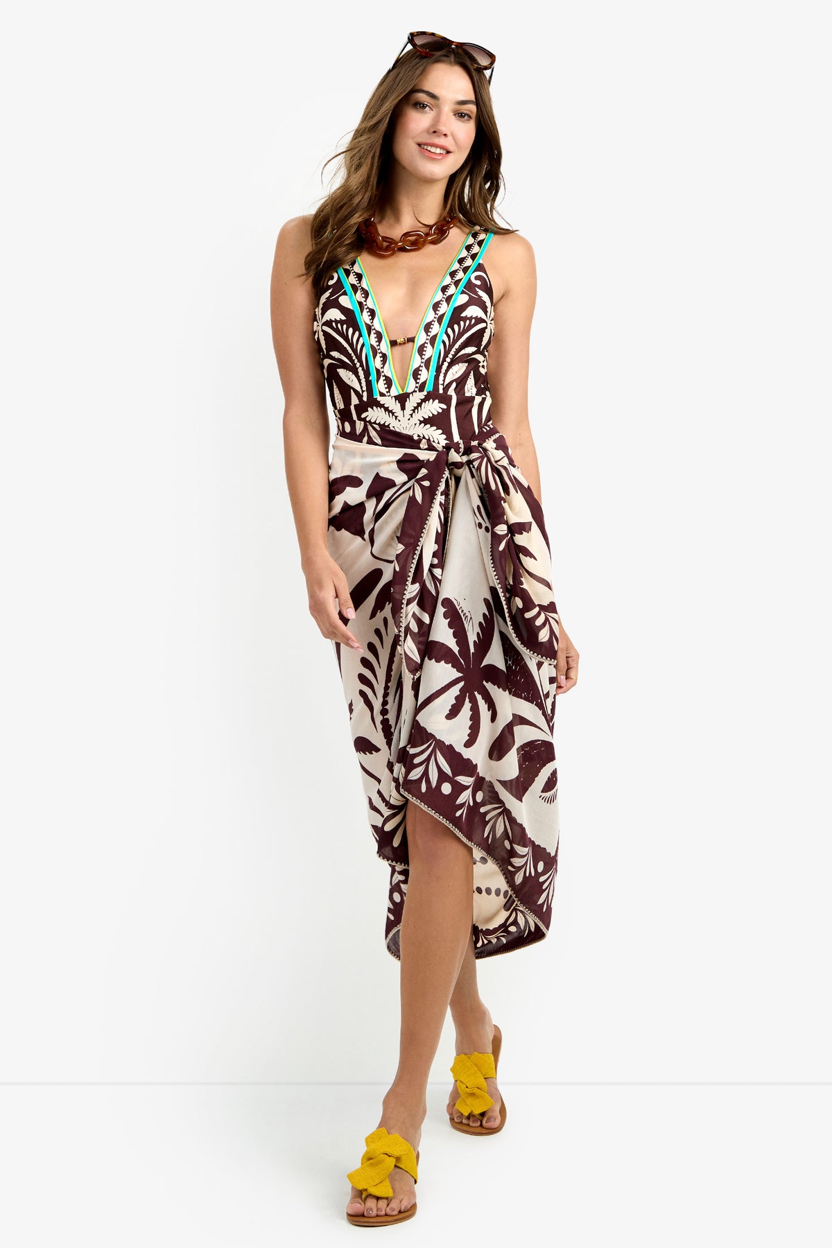 A woman stands and smiles against a white background, wearing the Pareo Swim Cover-Up—a sleeveless, deep V-neck dress with a bold brown and cream tropical print—paired with yellow sandals, a chunky necklace, and sunglasses on her head.