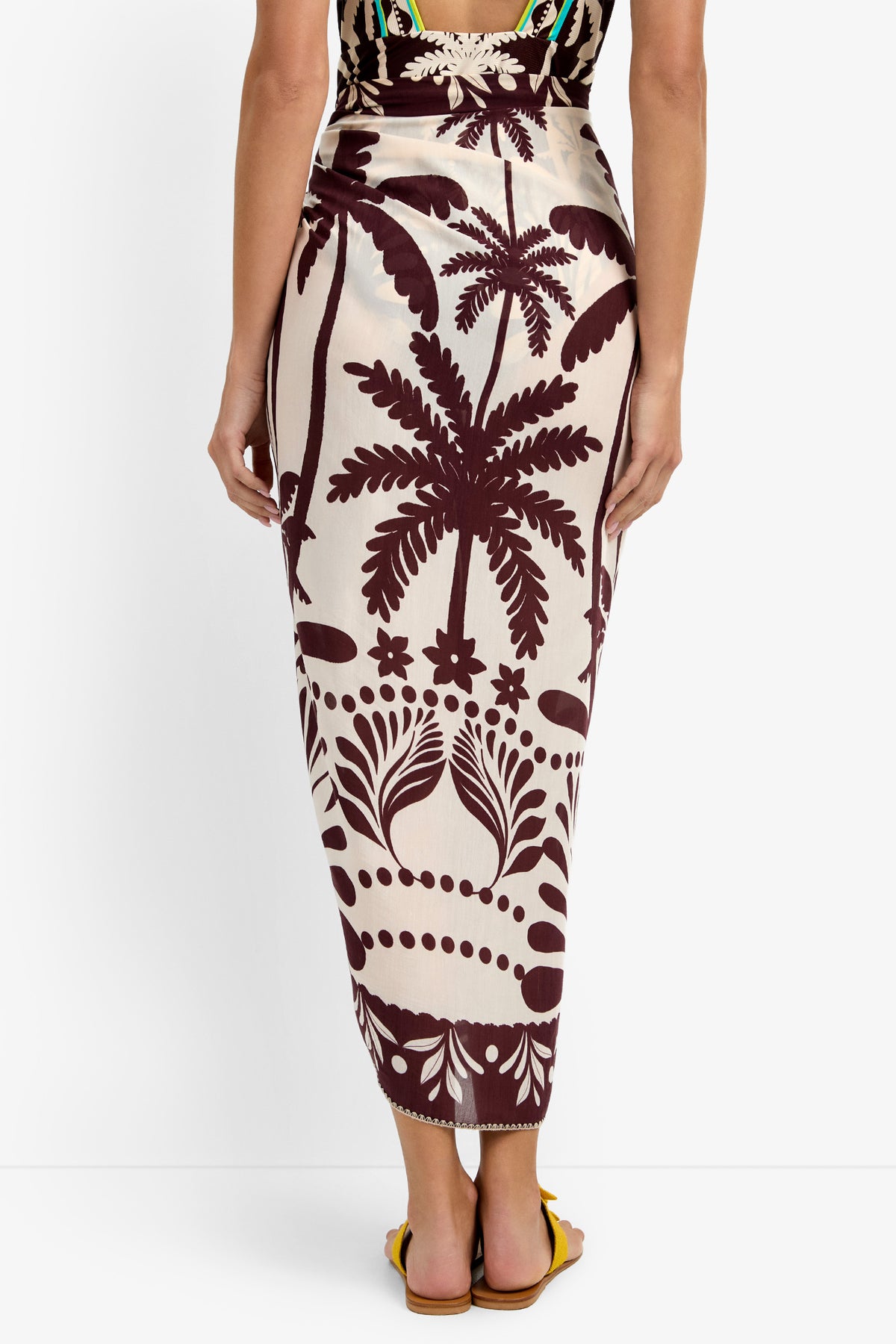 A woman seen from the back wears the Pareo Swim Cover-Up, a high-waisted, cream midi skirt featuring a bold maroon tropical palm and floral print. She pairs it with yellow sandals.
