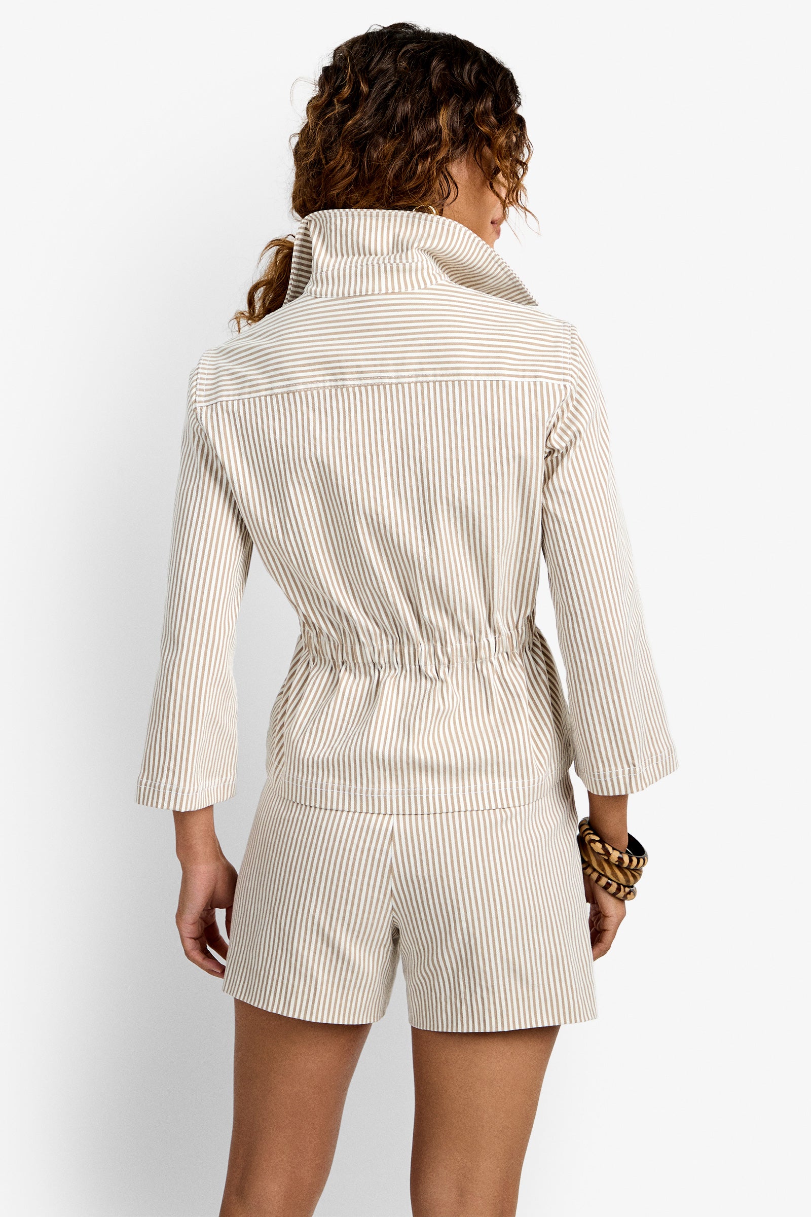 A woman with curly hair, shown from the back, wears the light-colored Rafferty Jacket with thin vertical stripes and matching shorts. She accessorizes with a large chunky bracelet on her wrist.
